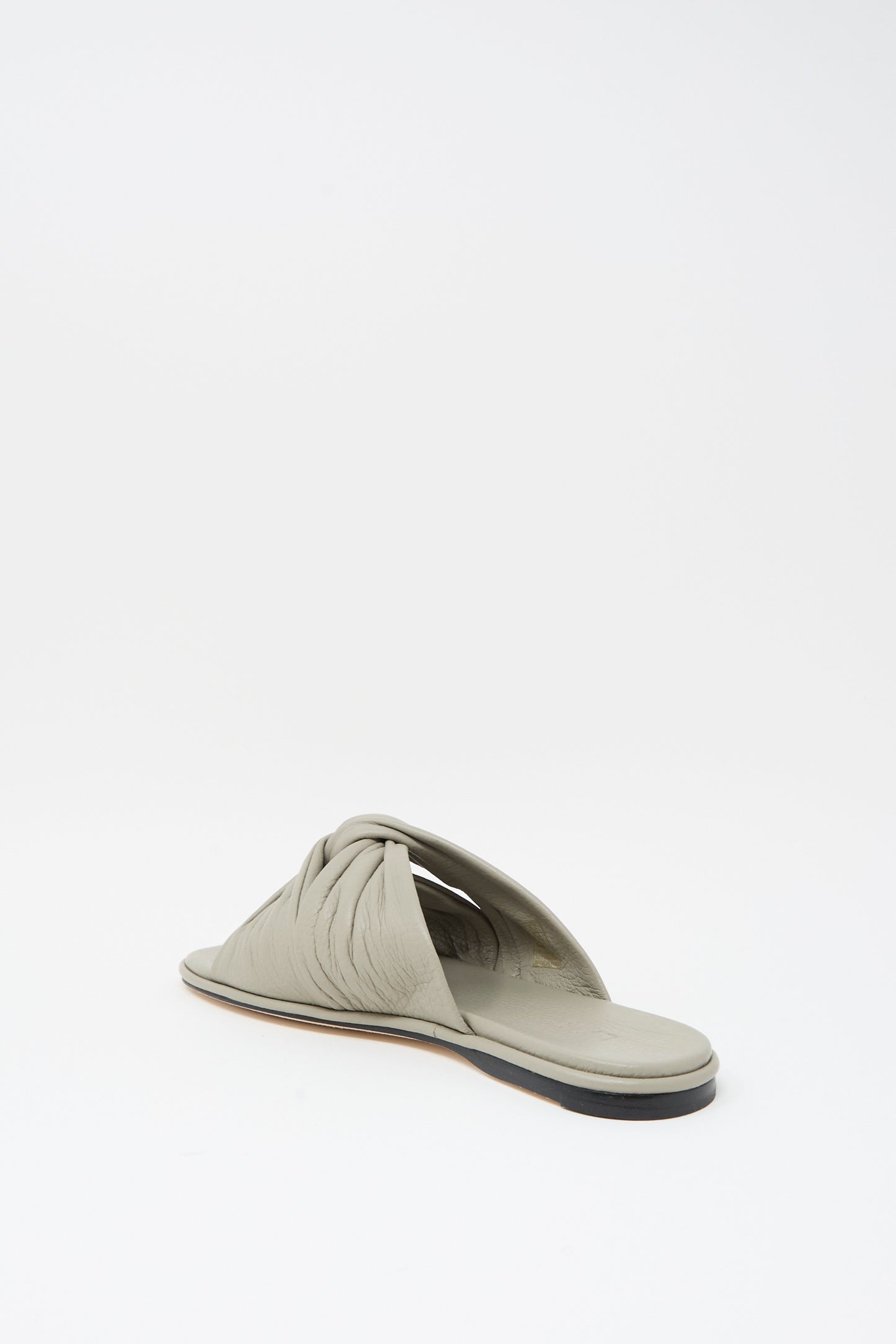 The Lauren Manoogian Twist Slide in Stone is a handmade light gray leather sandal featuring a twisted strap and flat sole, shown on a plain white background.