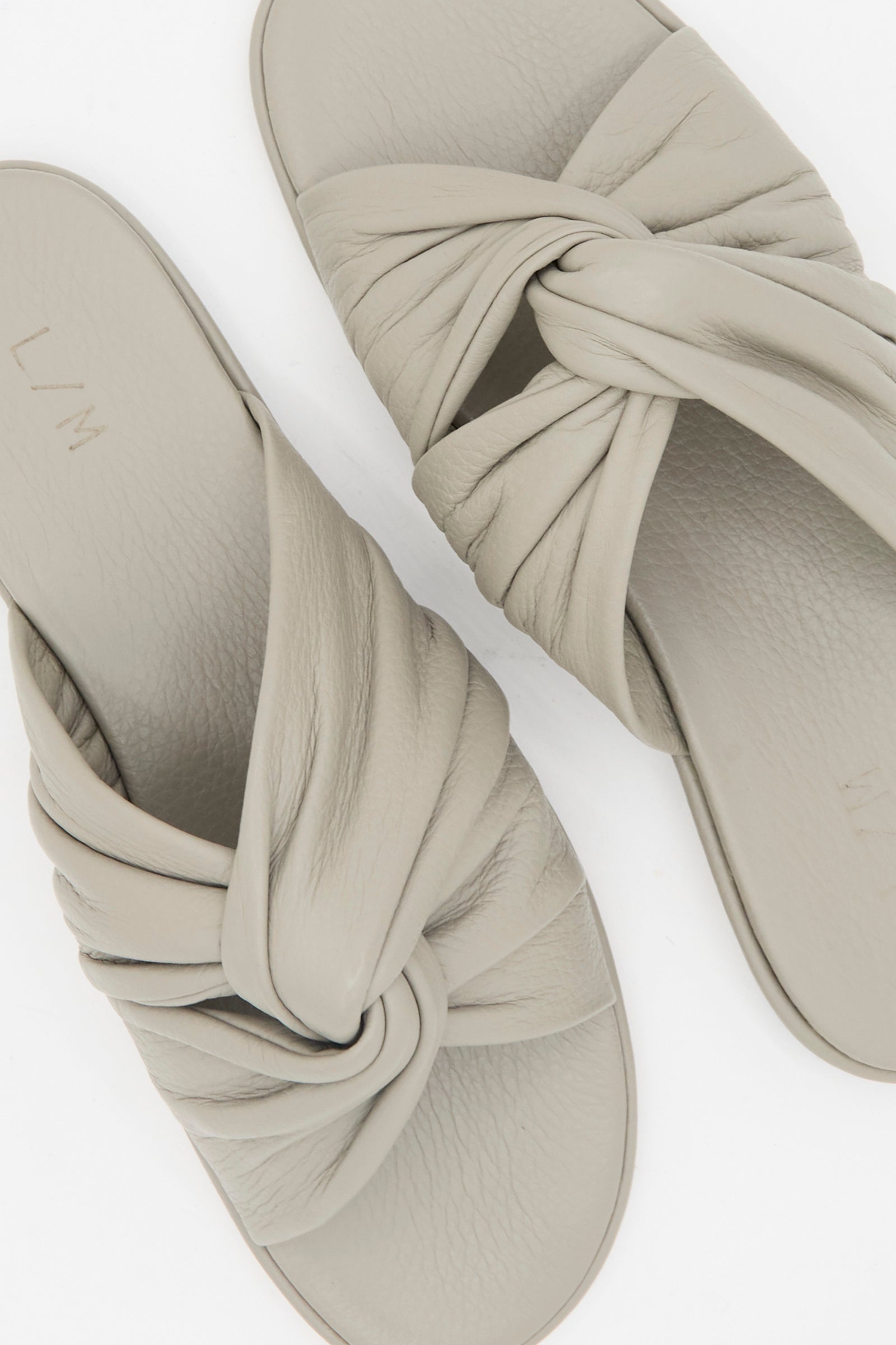 The Lauren Manoogian Twist Slide in Stone features light beige leather twist vamp straps and flat soles, shown against a white background.