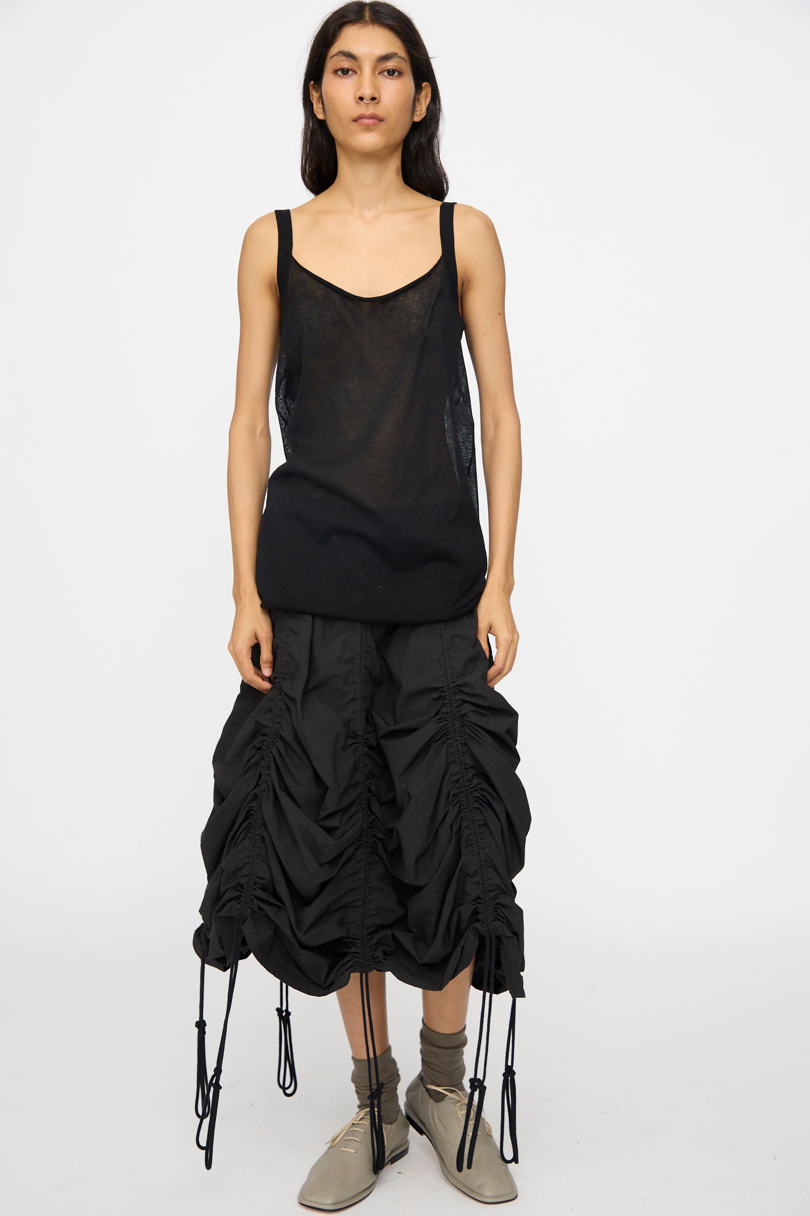 A person stands facing forward against a plain white background, wearing the Lauren Manoogian Veil Camisole in Black, a ruched black skirt with drawstrings, and beige ankle boots.
