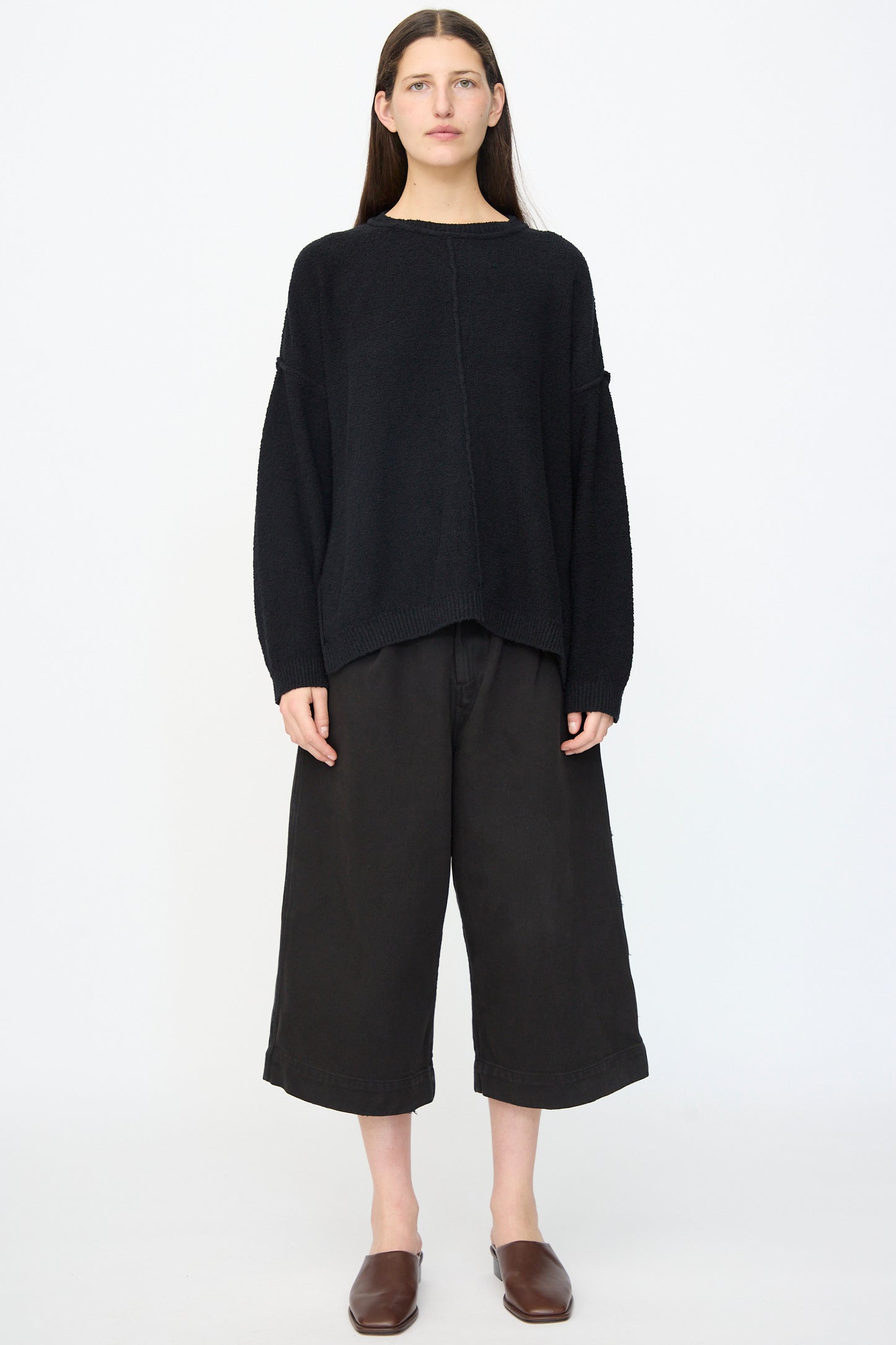 A person stands facing forward in a loose black sweater, Lauren Manoogian Washed Crop Trouser in Black, and brown slip-on shoes against a plain white background.