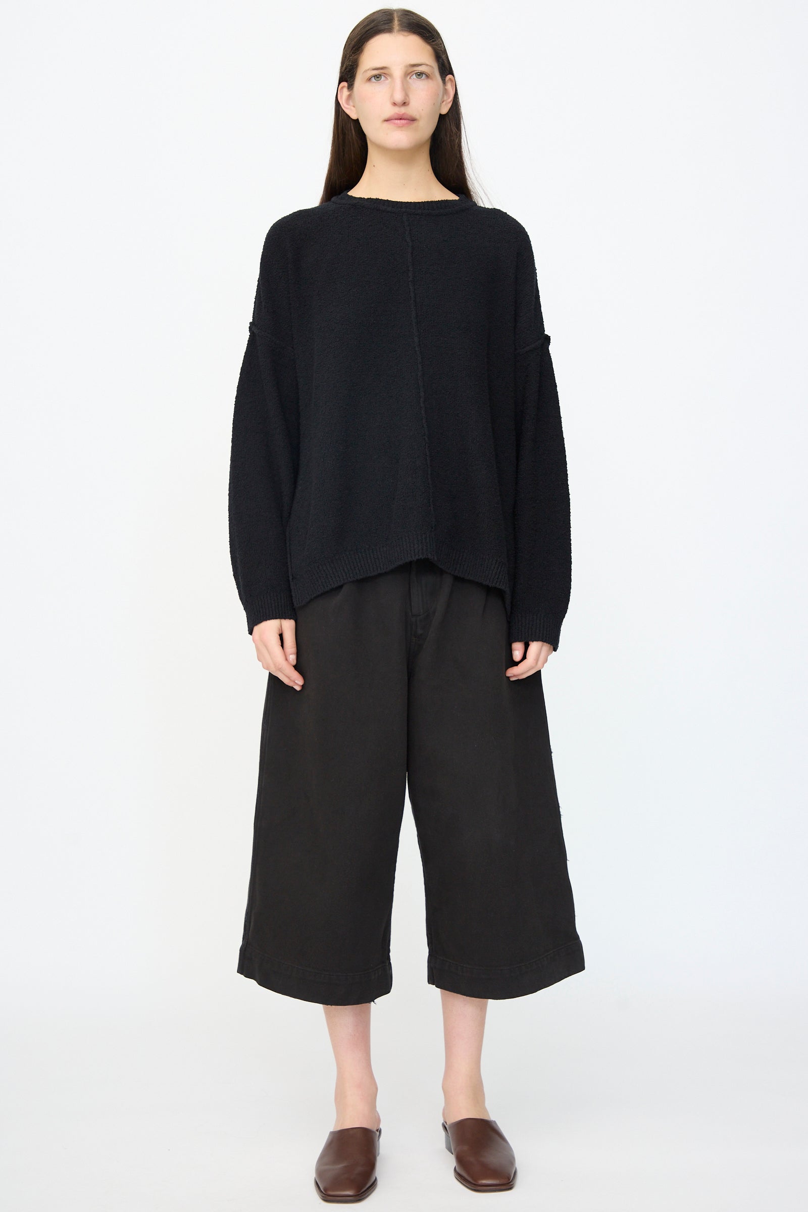 A person stands facing forward in a loose black sweater, Lauren Manoogian Washed Crop Trouser in Black, and brown slip-on shoes against a plain white background.
