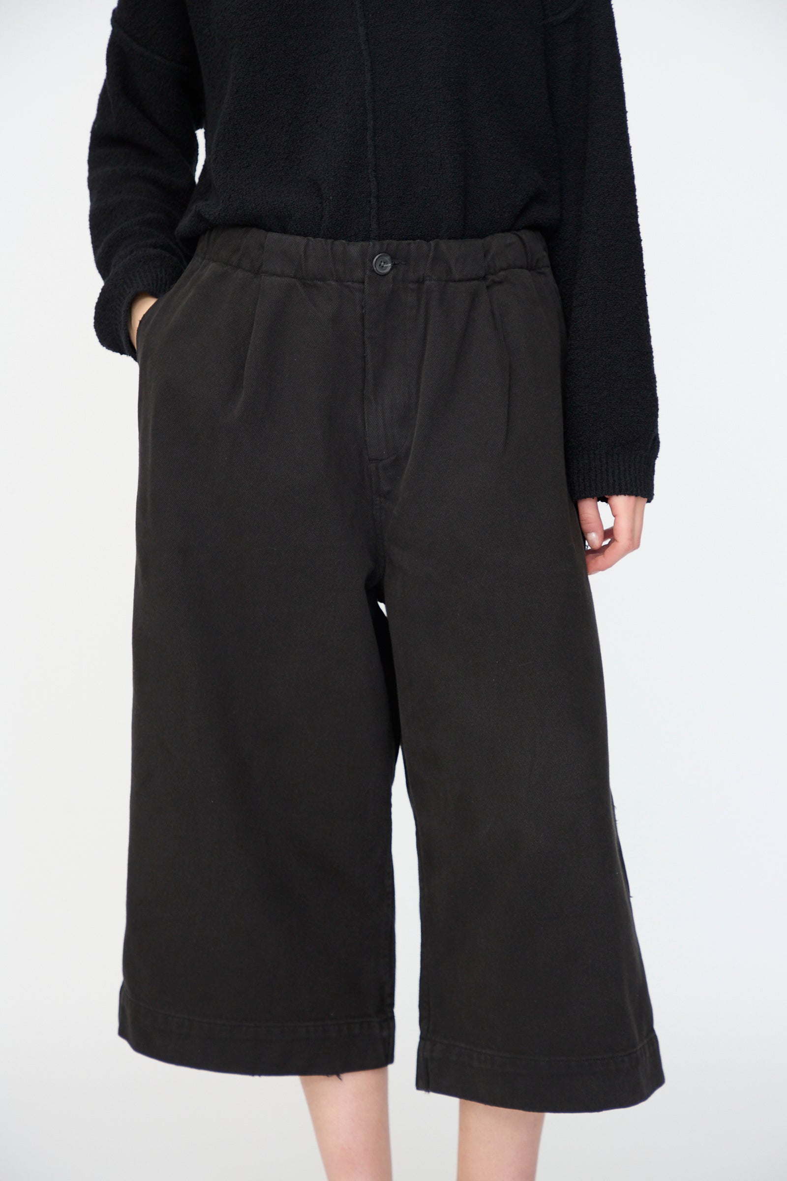 A person wearing a black long-sleeve top and Lauren Manoogian's Washed Crop Trouser in Black, featuring a relaxed fit and wide-leg cropped design, stands with hands in pockets against a plain white background.