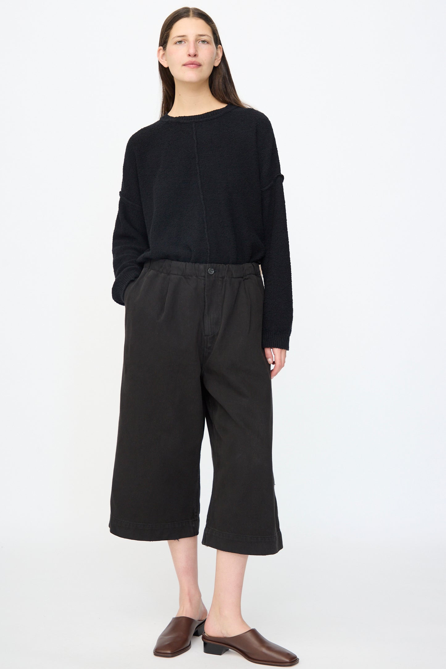 A person stands against a plain background wearing a relaxed fit black long-sleeve top, Lauren Manoogian Washed Crop Trouser in Black, and brown slip-on shoes.