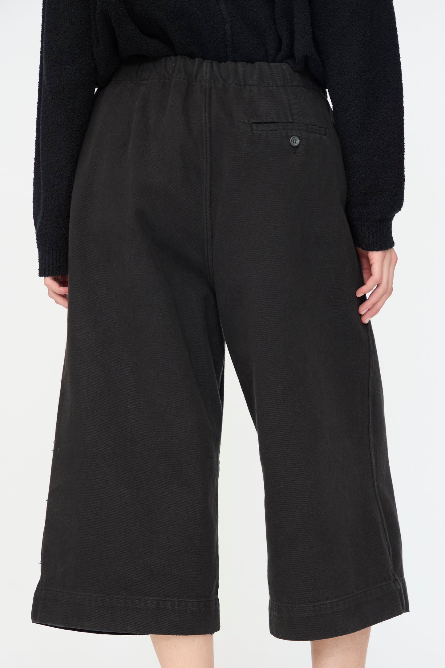 Model wearing Lauren Manoogian's Washed Crop Trouser in Black—relaxed fit cotton style with an elastic waistband and back pocket, shown from behind against a plain backdrop.