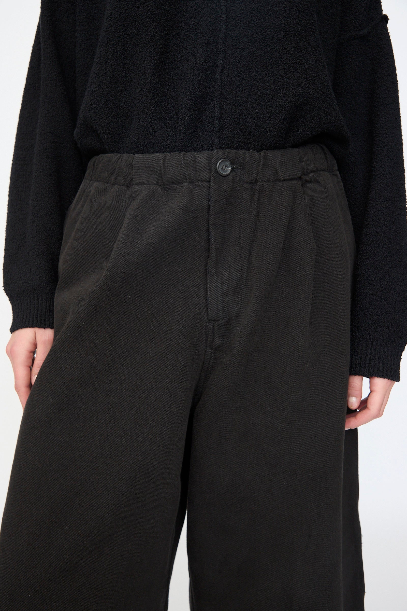 Model wears the Lauren Manoogian Washed Crop Trouser in Black, a relaxed-fit cotton pant with button and elastic waistband, paired with a black textured long-sleeve top. Shown from shoulders to knees.