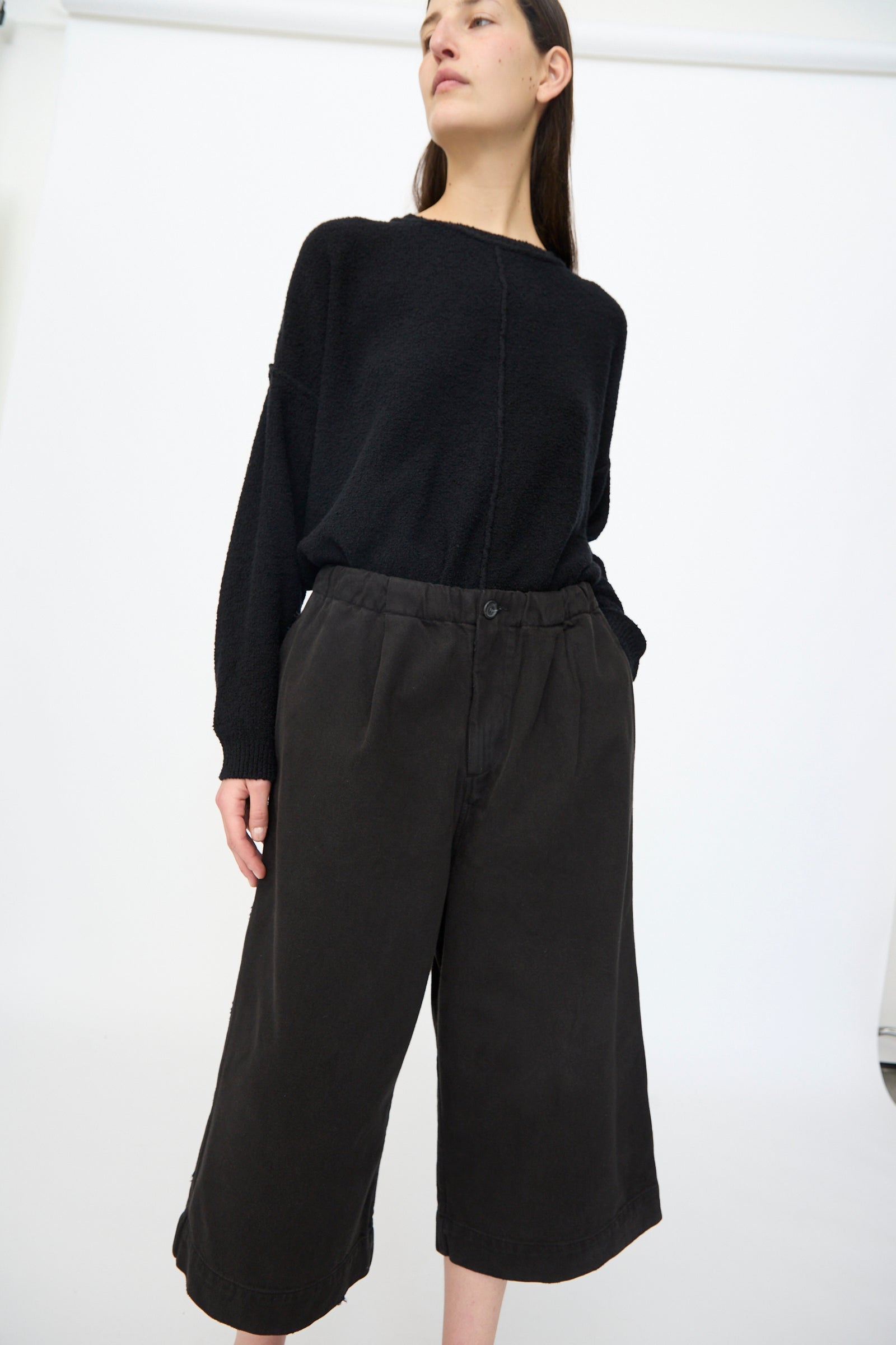A person stands at an angle against a white background, wearing Lauren Manoogian’s Washed Crop Trouser in Black paired with a loose black long-sleeve top.