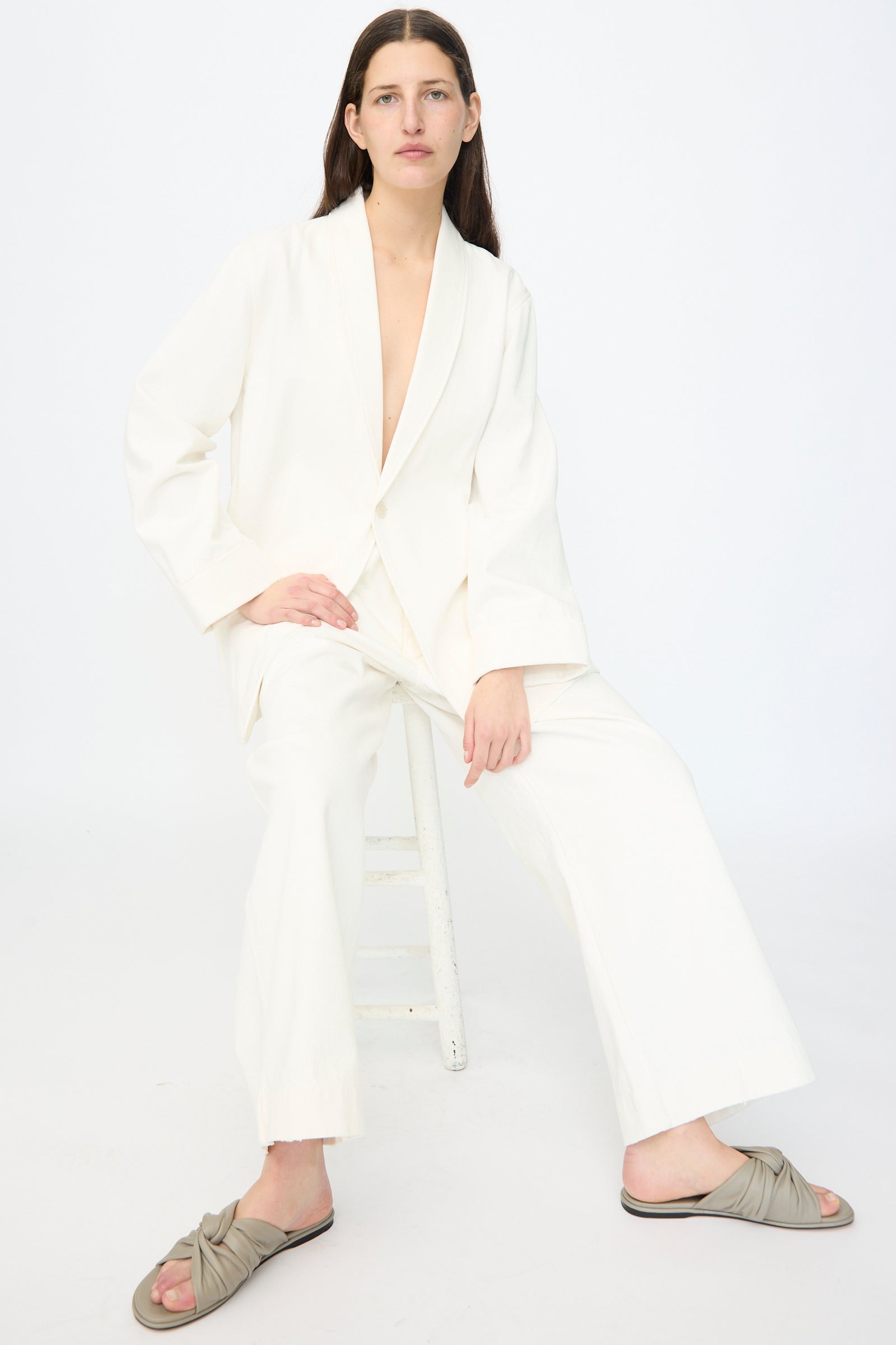 A person with long brown hair sits on a white stool, wearing the Lauren Manoogian Washed Jacket in White and beige slide sandals, against a plain white background.