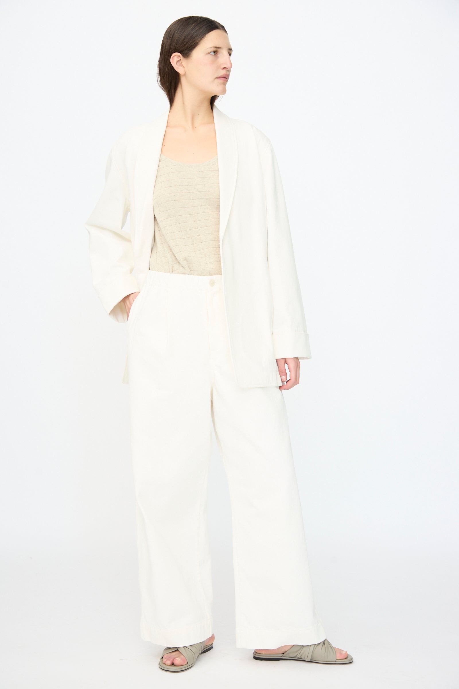A woman stands against a plain background wearing Lauren Manoogian’s Washed Trouser in White—a relaxed, elastic-waist style—paired with a loose white blazer, beige top, and gray sandals.