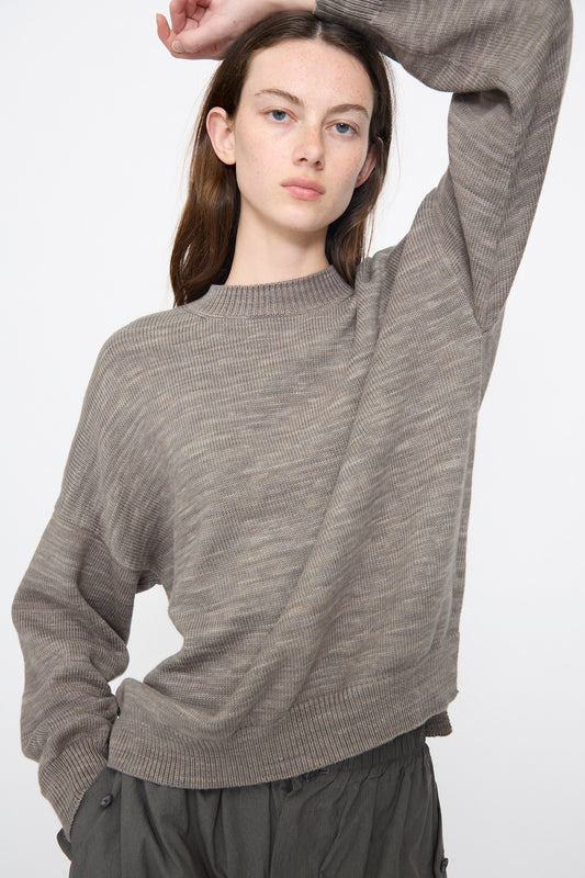 A woman with long brown hair wears the Lauren Manoogian Watercolor Crewneck in Rock, a relaxed-fit taupe sweater, paired with dark gray pants, her arm raised behind her head.