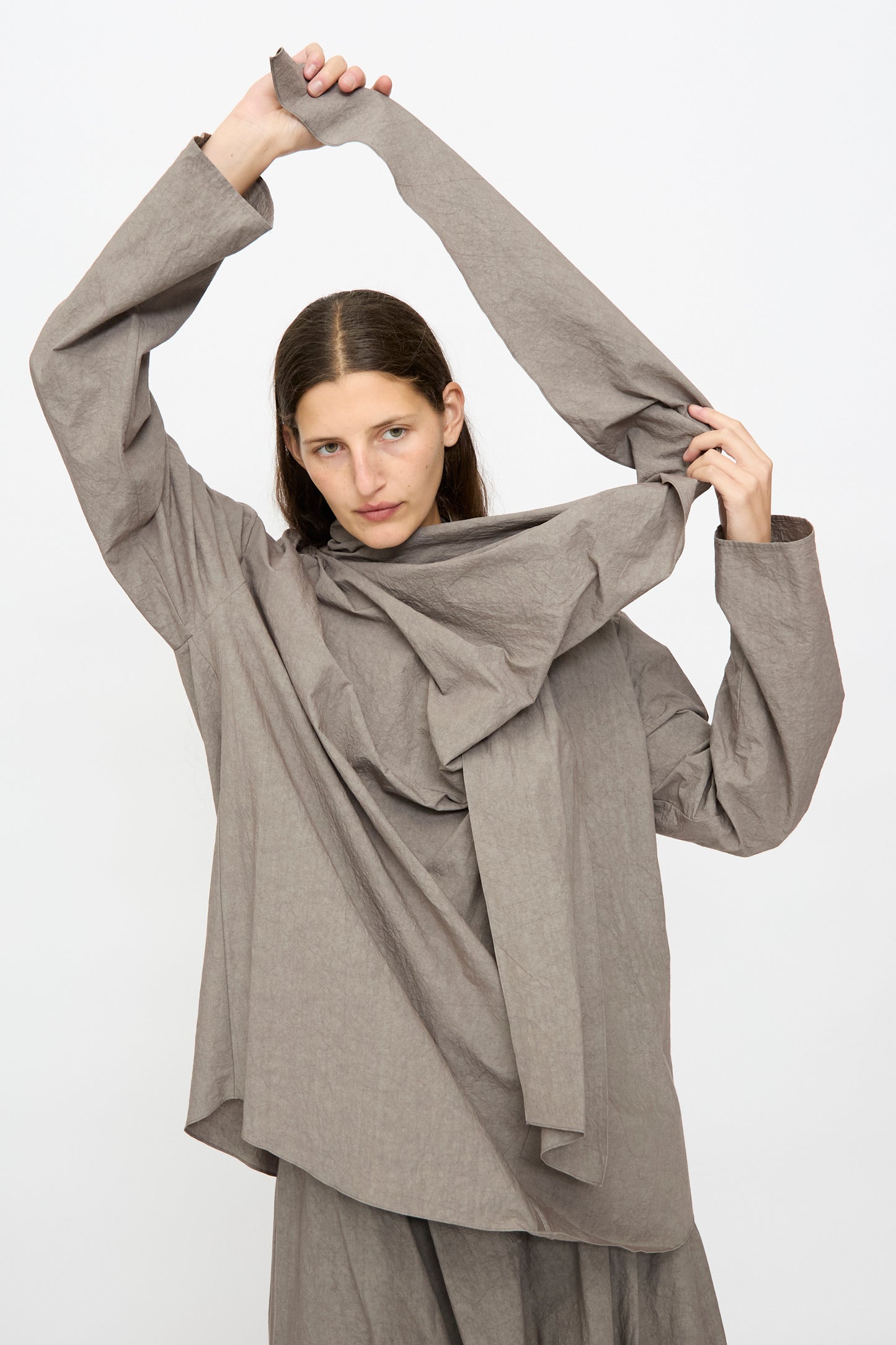 A person wearing the Lauren Manoogian Wrap Top in Cinder stands against a plain background, holding one sleeve above their head with a neutral expression.