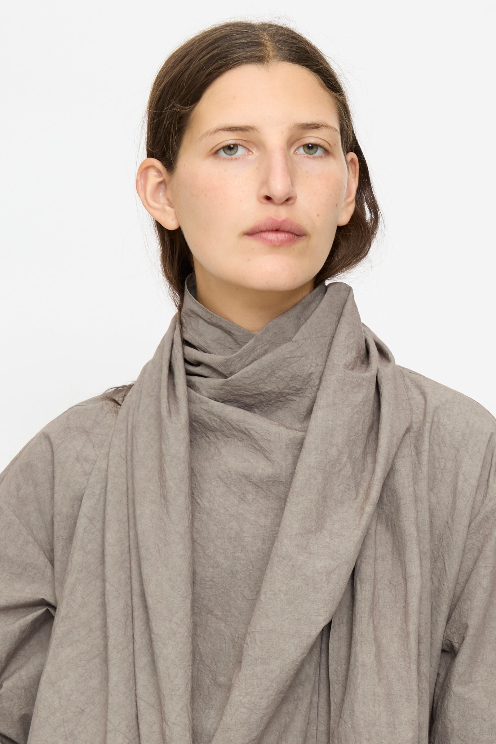 A person with long brown hair wears the Lauren Manoogian Wrap Top in Cinder, featuring a loose, draped silhouette and long sleeves, standing against a plain light background.