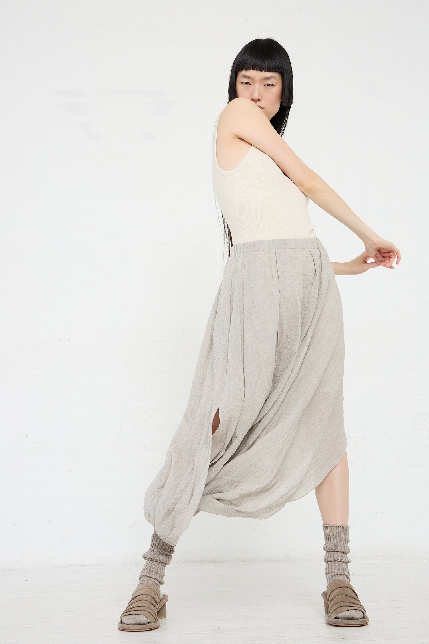 A woman with black hair in a beige cotton/linen blend tank top and draped grey pants poses gracefully against a white background wearing the Gauze Twist Skirt in Ash by Lauren Manoogian.