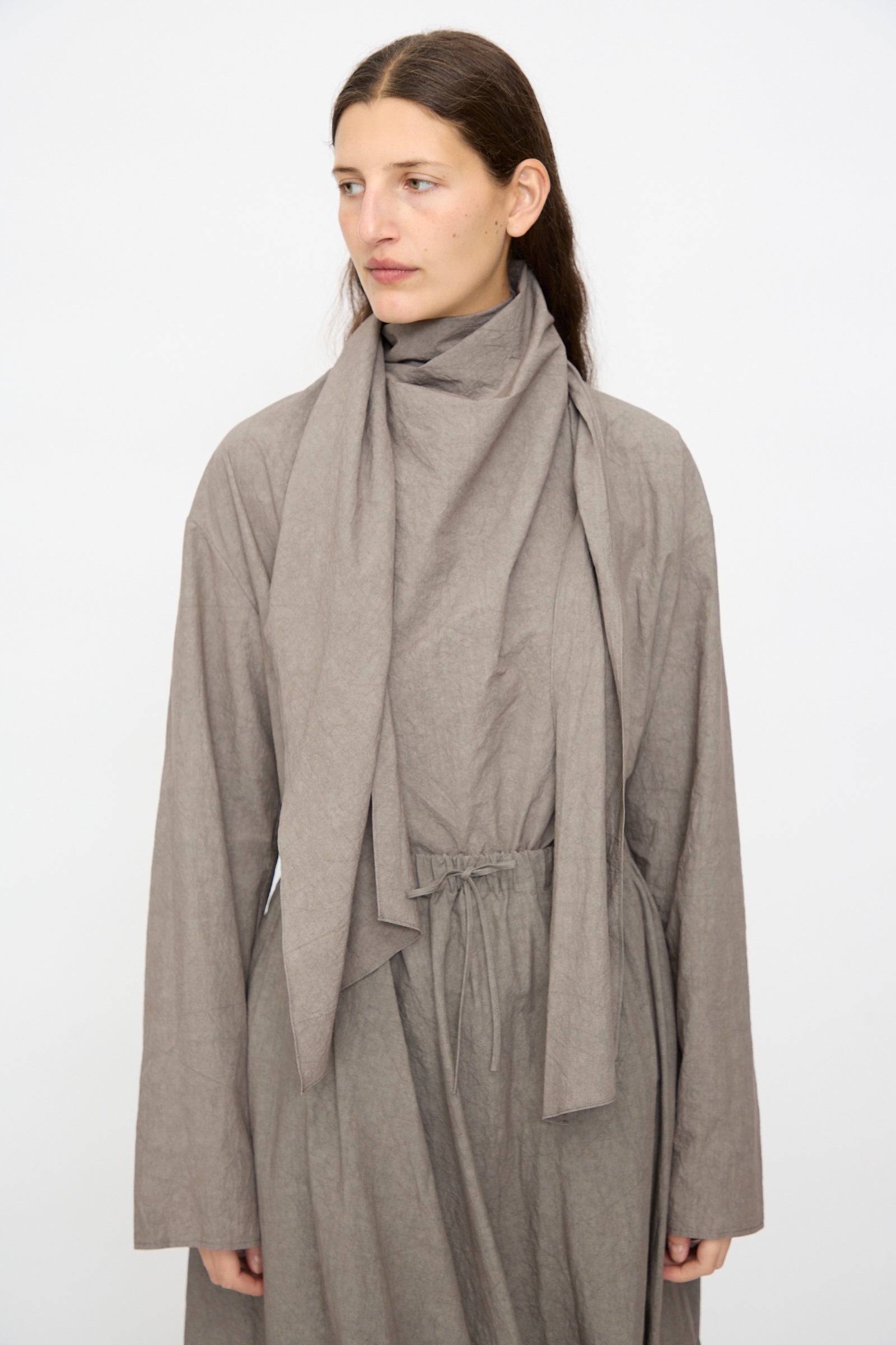 A woman with long brown hair wears the Lauren Manoogian Wrap Top in Cinder, a loose, light brown cotton long sleeve piece with matching scarf and drawstring waist, standing against a plain white background.