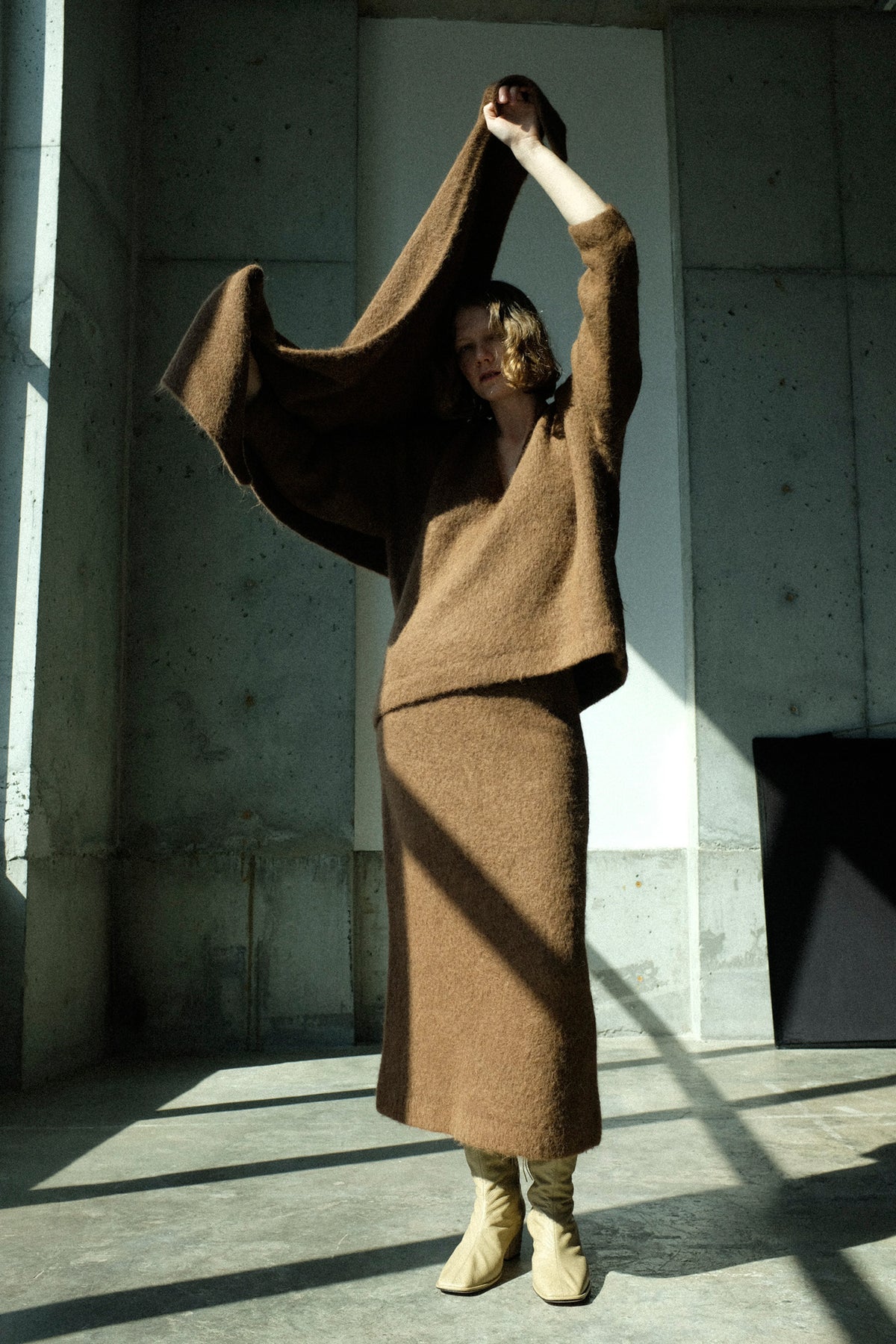 A model in a minimalist concrete room wearing Lauren Manoogian's Brushed Pencil Skirt in Rust, and Brushed V Neck in Rust, holding the Brushed Triangle Wrap in Rust open with both arms raised above their head.
