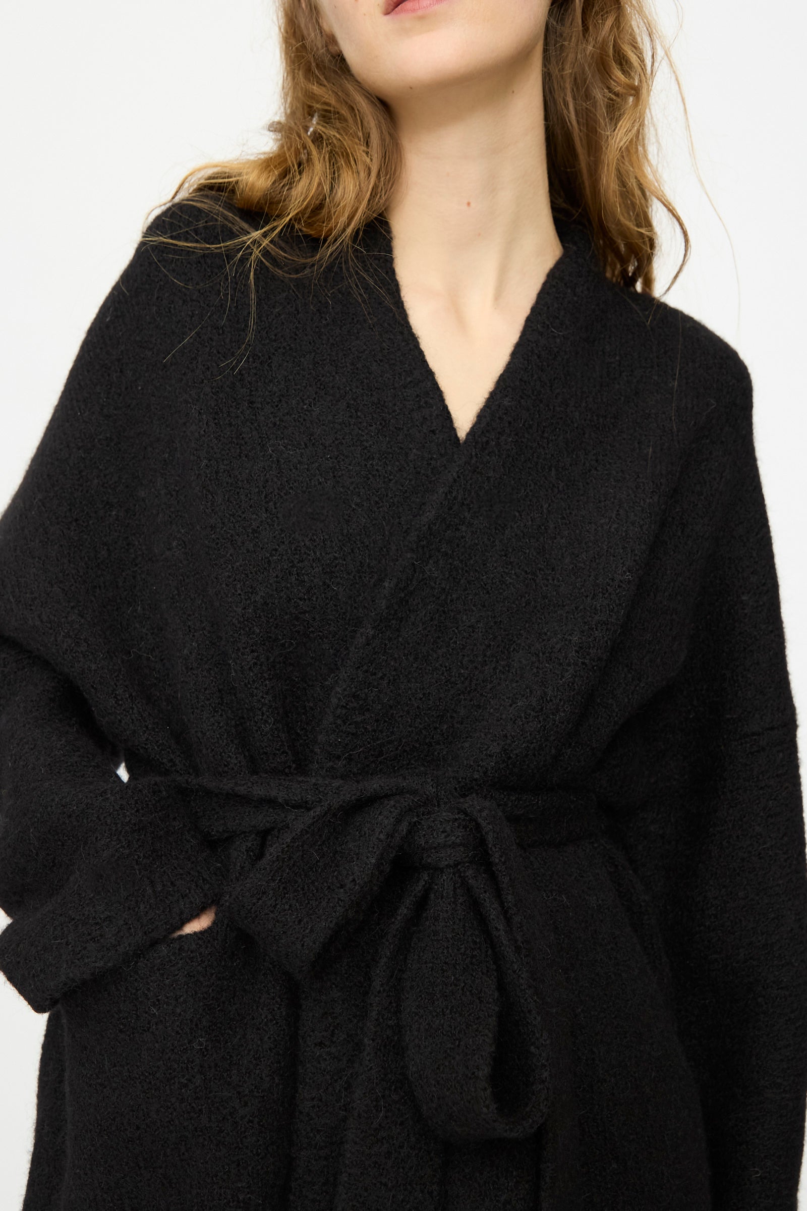 A person wears the Lauren Manoogian Coto Cardigan in Black, a hand-loomed alpaca piece with a tie belt, made in Peru, standing against a plain white background.