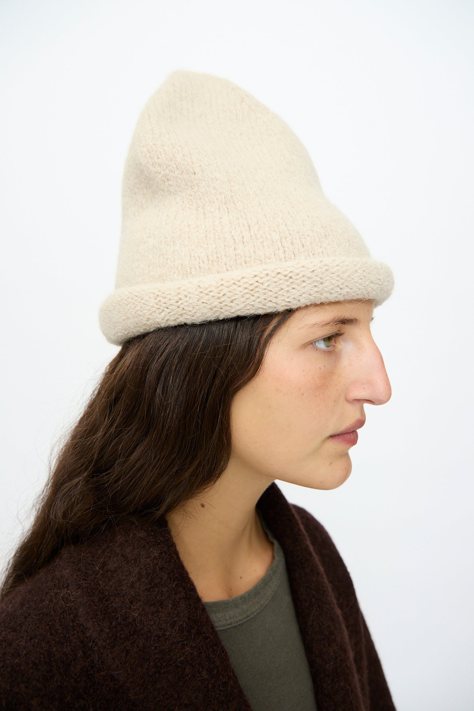 A person with long brown hair wears the Lauren Manoogian Crown Beanie in Antique Melange and a dark brown coat, shown in profile against a plain light background.