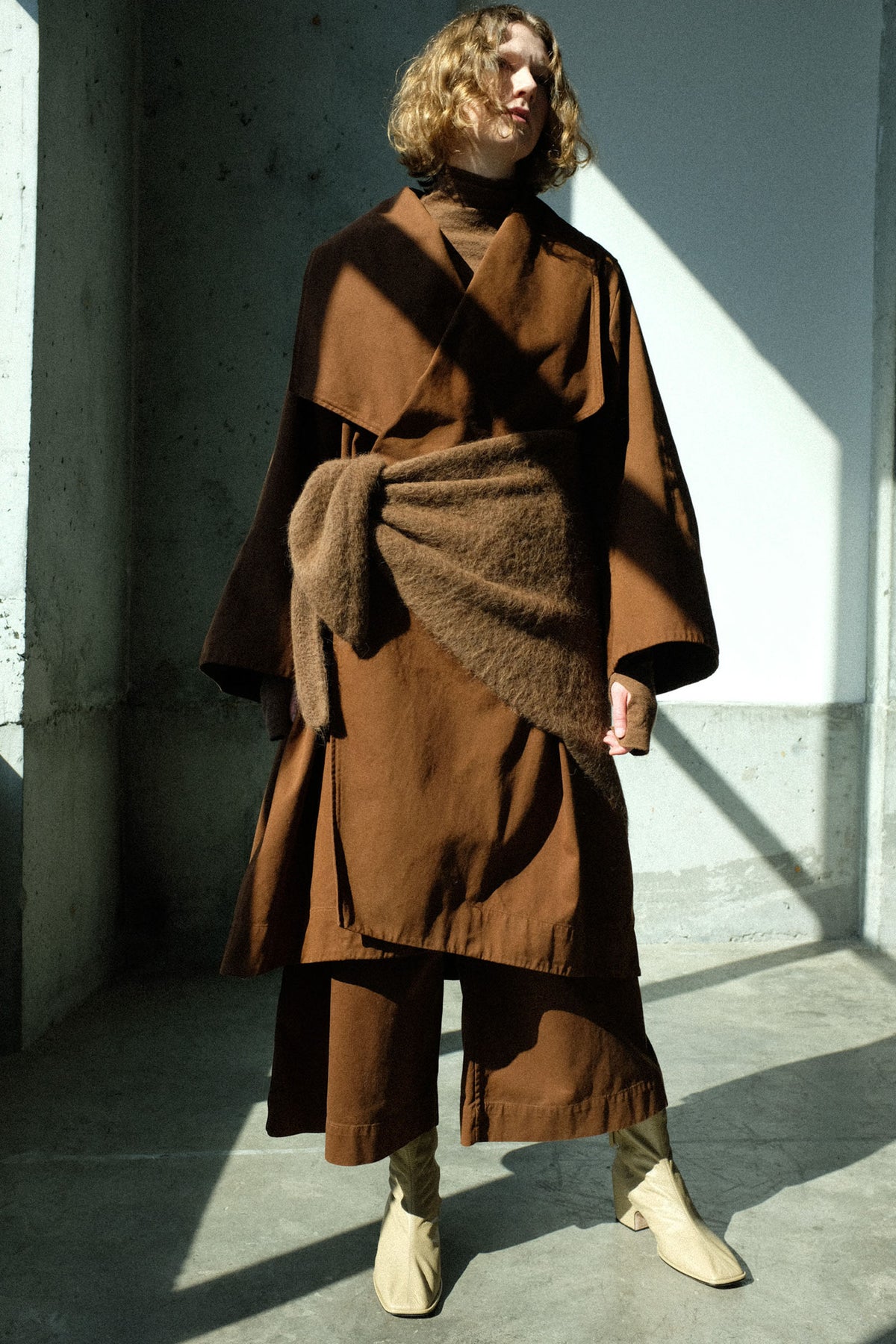 A model wearing pieces from Lauren Manoogian's FW26 collection standing in a concrete room with dramatic shadows.
