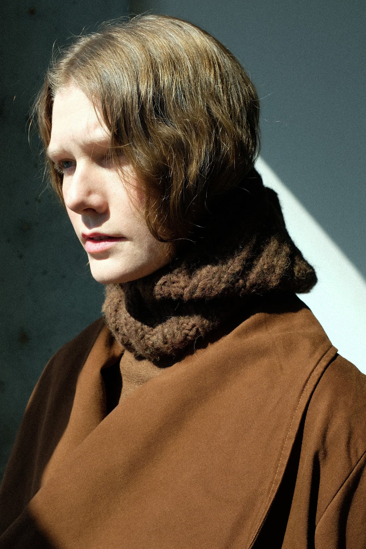 A person shown in profile wearing Lauren Manoogian's Handknit Rib Balaclava in Rust Melange around her neck, photographed against a dark gray wall with strong sunlight and shadow.