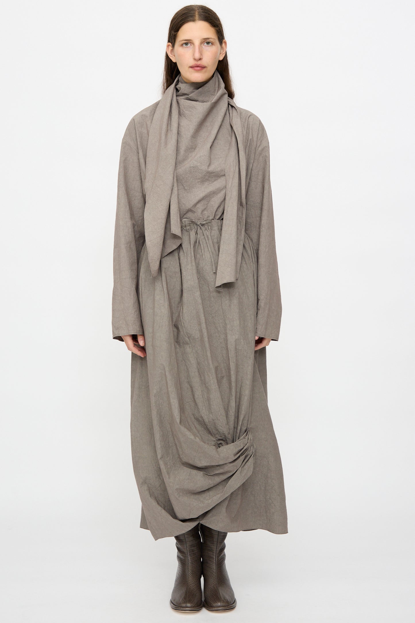 A woman faces forward against a plain white background, wearing the Lauren Manoogian Wrap Top in Cinder styled with a draped scarf detail, gathered waist, and ankle boots.