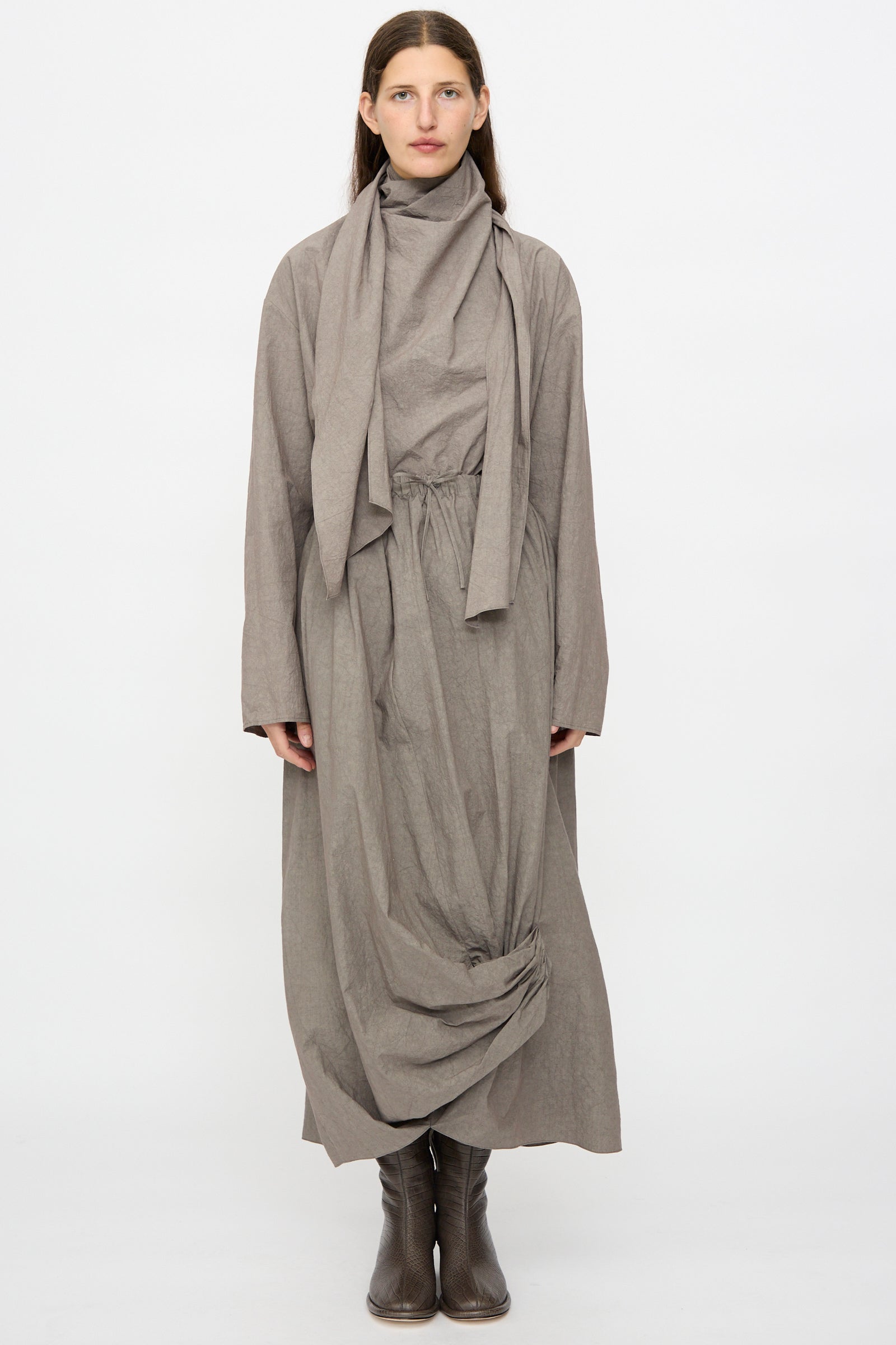 A woman faces forward against a plain white background, wearing the Lauren Manoogian Wrap Top in Cinder styled with a draped scarf detail, gathered waist, and ankle boots.