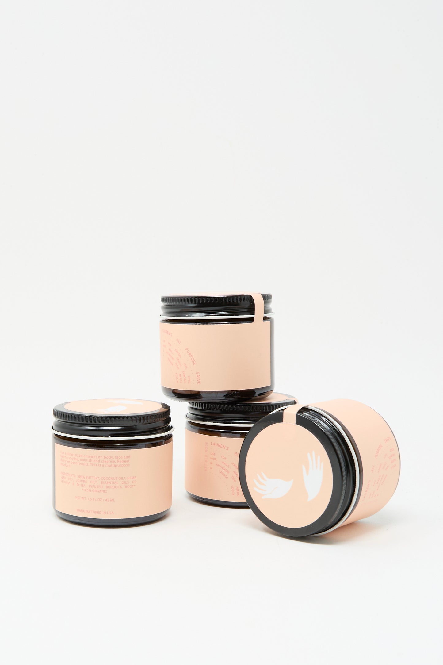 Three small 45ml dark glass jars of Lauren’s All Purpose Rose Vetiver Salve with peach labels and black lids, featuring a white winged logo, are arranged on a white background, suggesting premium vegan skincare.