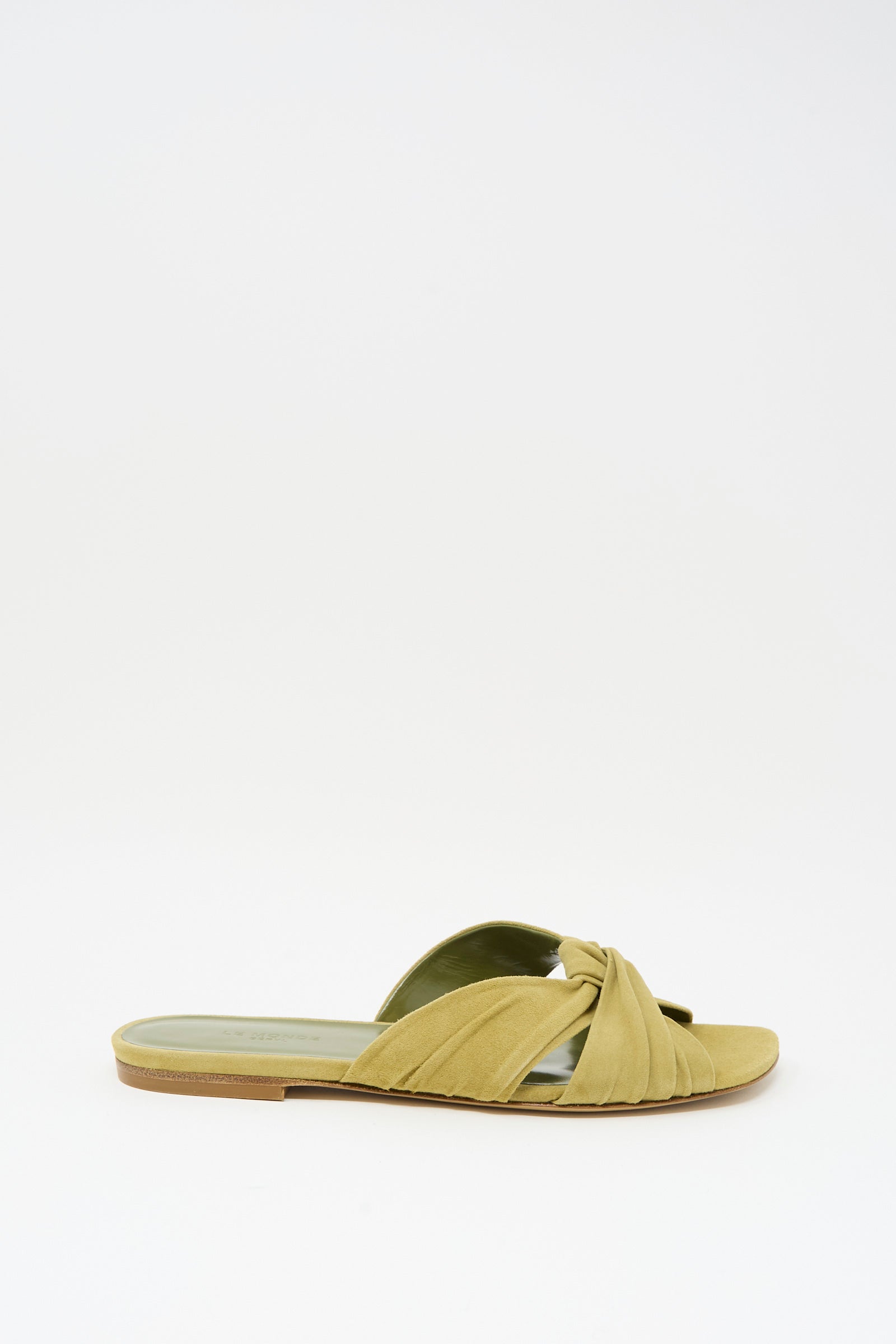 The Antonia Flat in Grey Green by Le Monde Beryl features a single light green suede slide with a twisted strap, open toe, and light brown sole, showcased on a plain white background.
