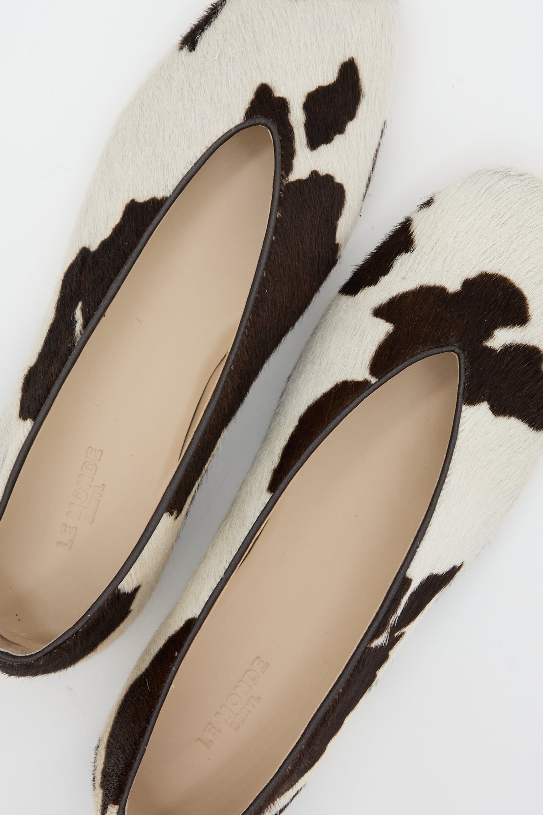 The Calf Hair Regency Slipper in Cow by Le Monde Beryl offers a stylish slip-on design with a black and white cow print pattern, enhanced with chic leather piping for an elegant finish.