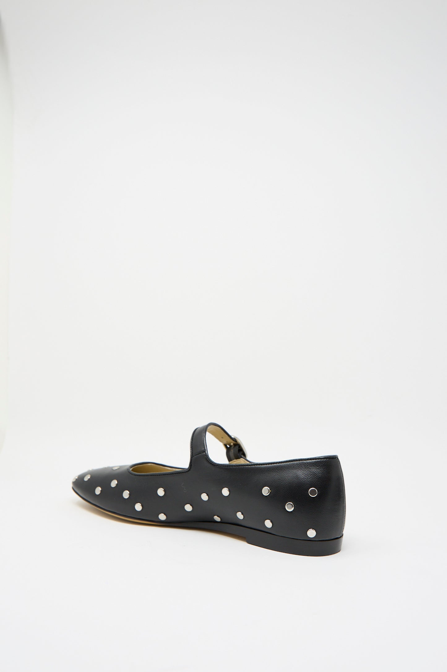 The Studded Leather Mary Jane in Black by Le Monde Beryl features a black leather flat design adorned with polka dots, a strap, and silver metal studs, all showcased from the back side on a plain white background. 