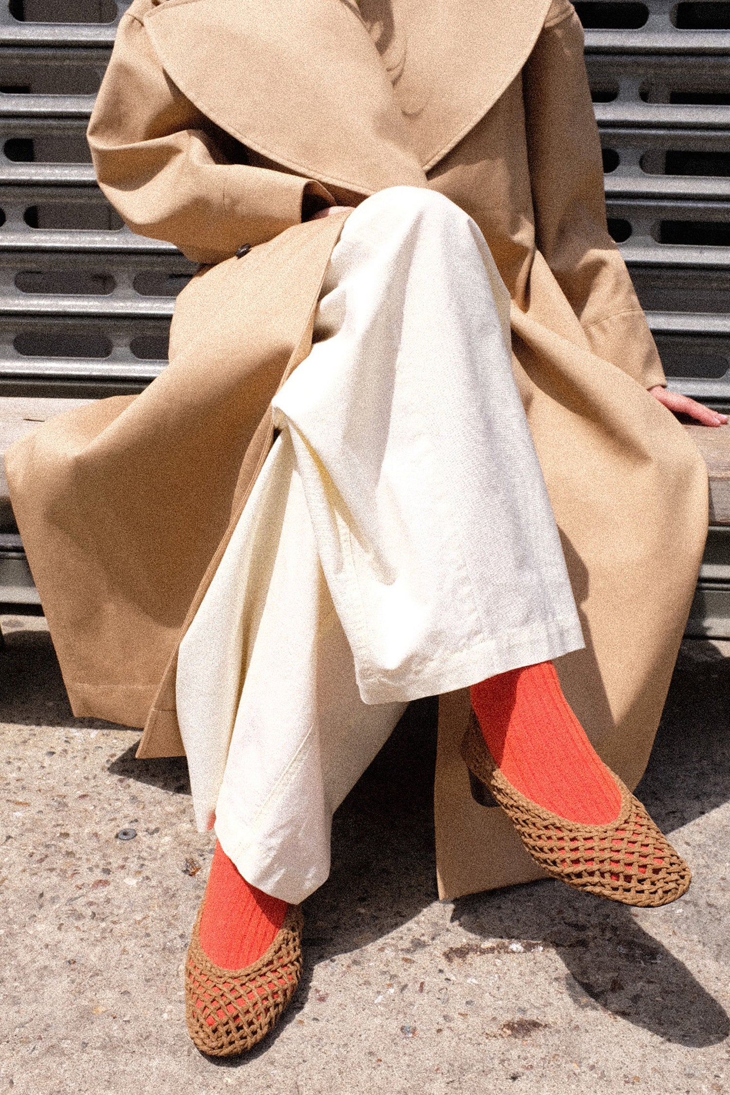 Person wearing a beige coat, white pants, and Le Monde Beryl Knotted Ballet Pump with orange sock, sitting on a bench.