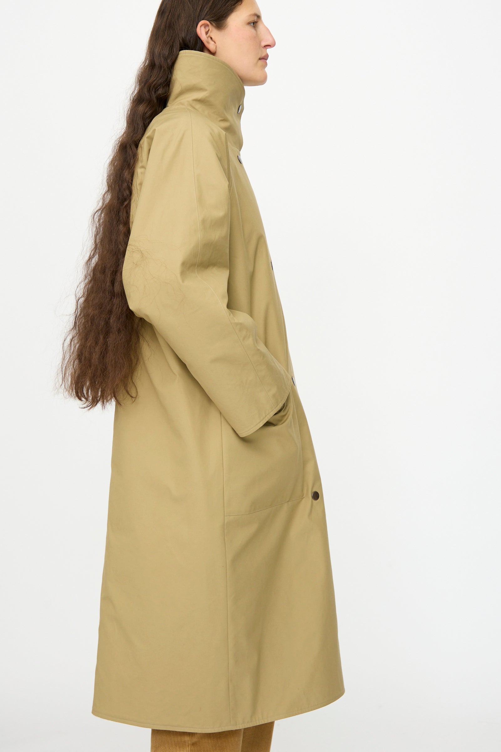 A person with long brown hair stands in profile, hands in pockets, wearing the Les Prairies De Paris Dickens Moon Reversible British Trench in Taupe against a plain white background.