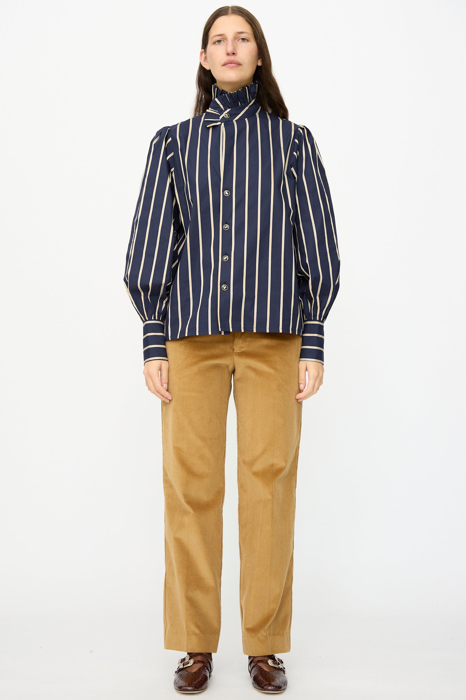 A person stands facing forward, wearing a navy and white vertical striped blouse, Gaoutcho Trouser in Miel by Les Prairies De Paris, and brown shoes, against a plain white background.