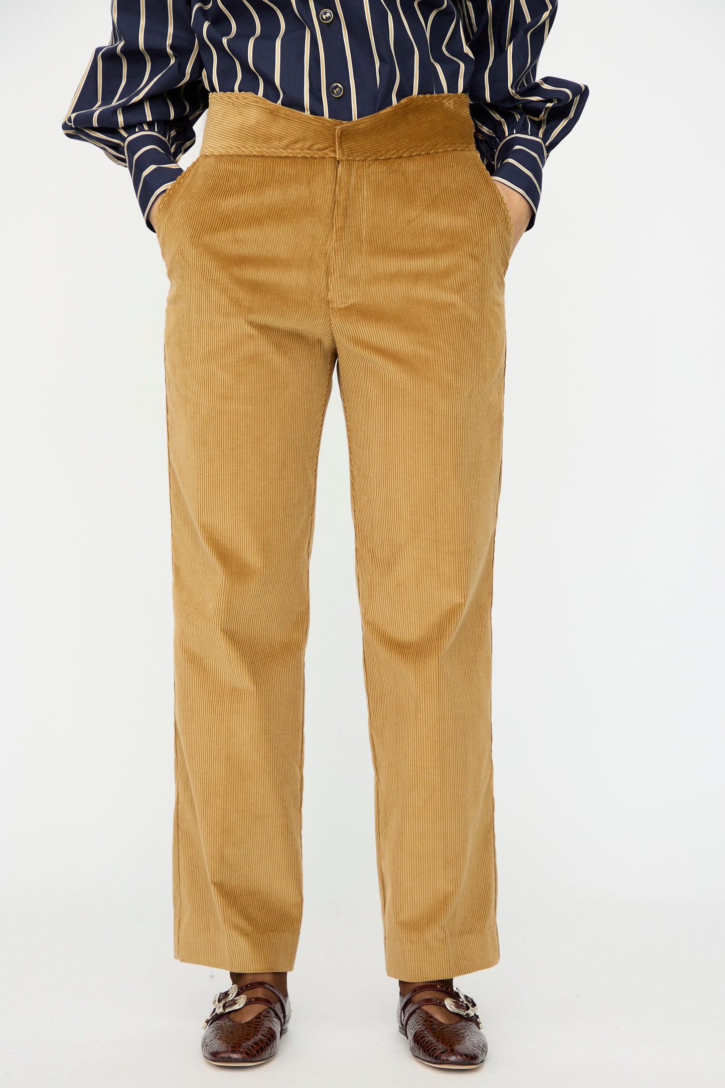 A person in Les Prairies De Paris' Gaoutcho Trouser in Miel, paired with a navy striped shirt and brown loafers, stands hands in pockets against a white background. The trousers' high waist adds classic French style.