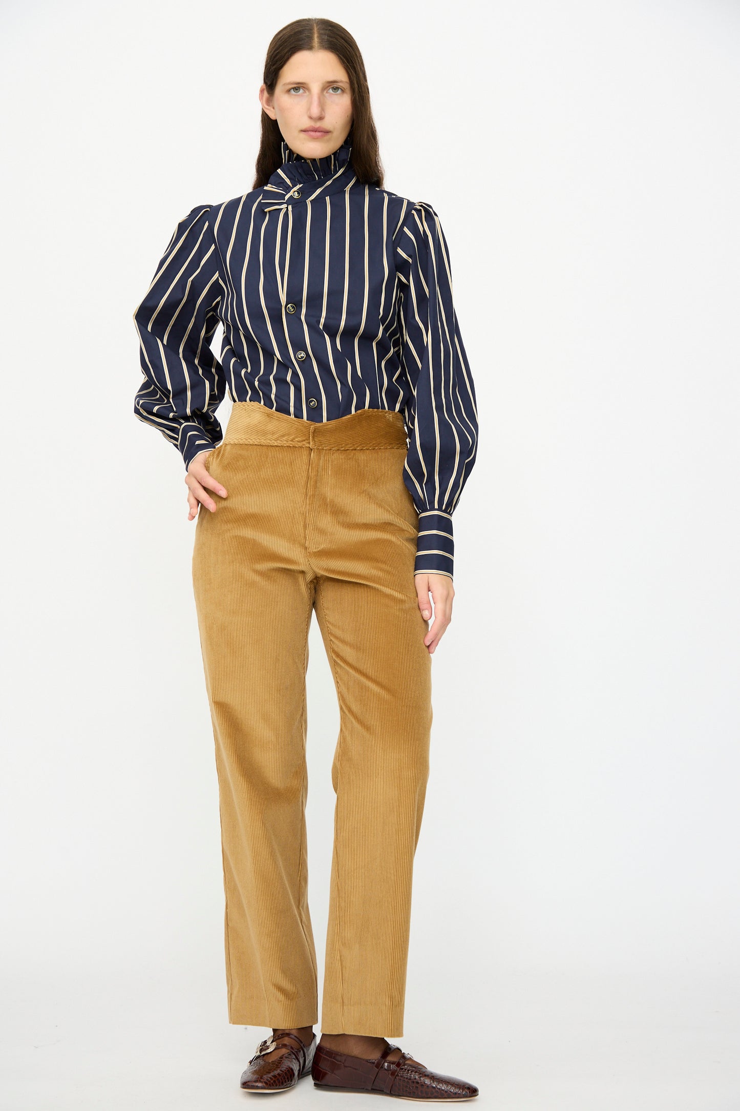 A person stands against a plain background wearing a navy striped button-up shirt, Les Prairies De Paris Gaoutcho Trouser in Miel, and brown shoes.
