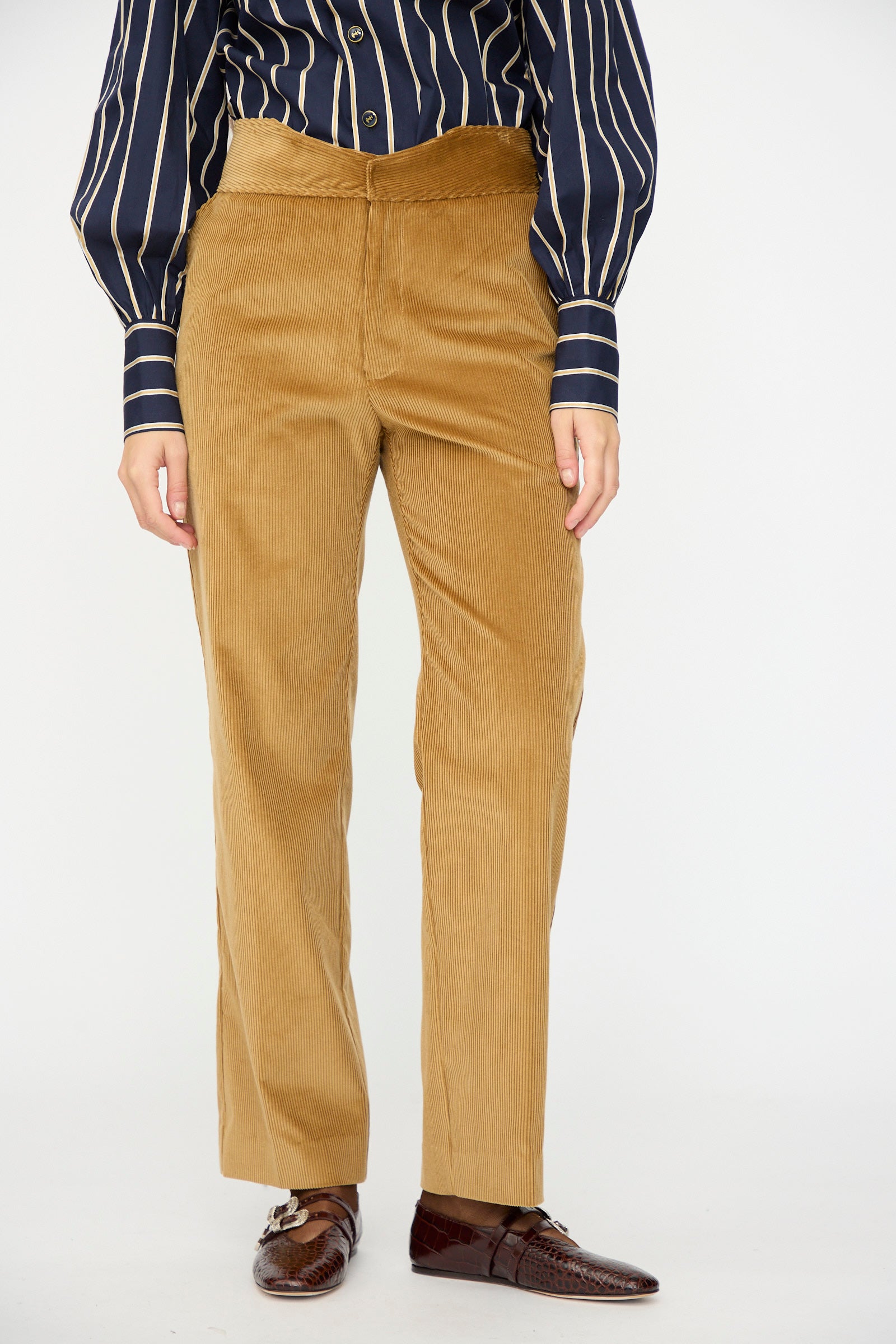 Person wearing Les Prairies De Paris Gaoutcho Trouser in Miel, a navy shirt with white stripes, and brown dress shoes, standing against a plain background.