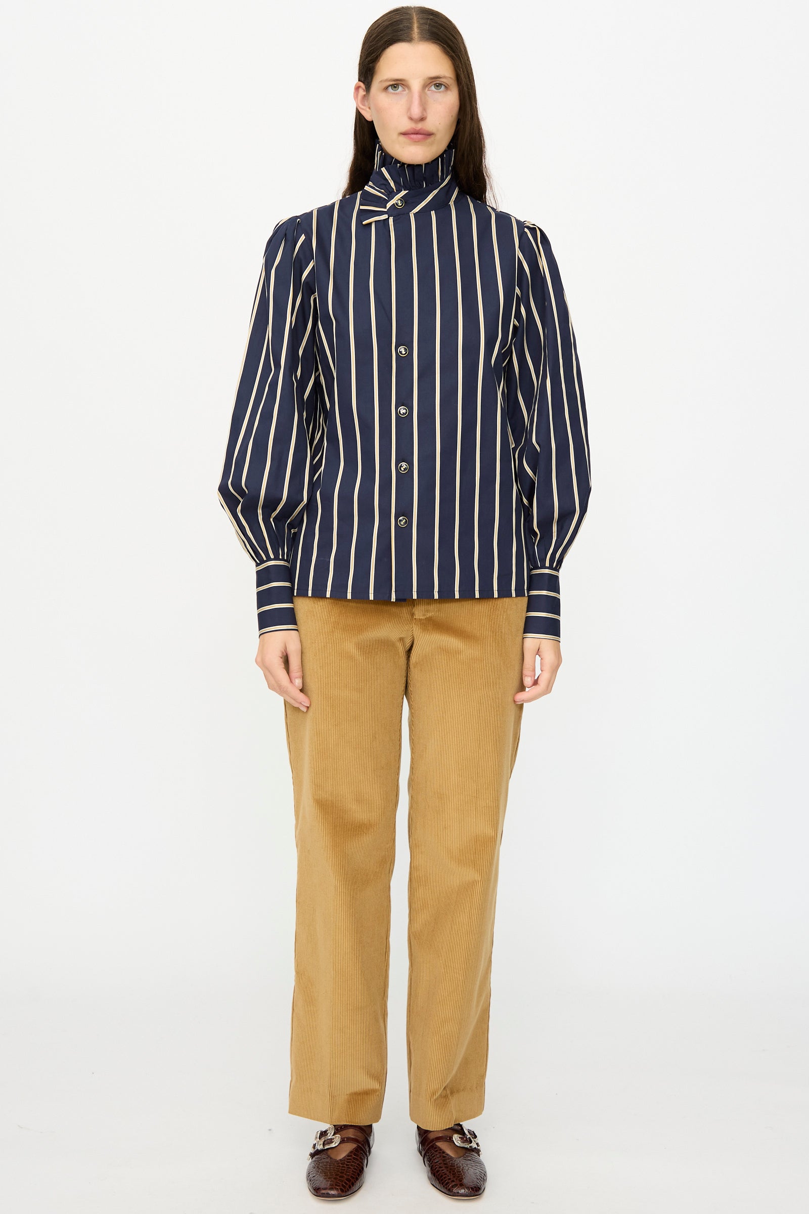 A person stands facing forward wearing the Les Prairies De Paris Cotton Josephine Shirt in R Studio, featuring navy blue and white stripes with a ruffled collar, paired with tan pants and brown shoes against a plain white background.