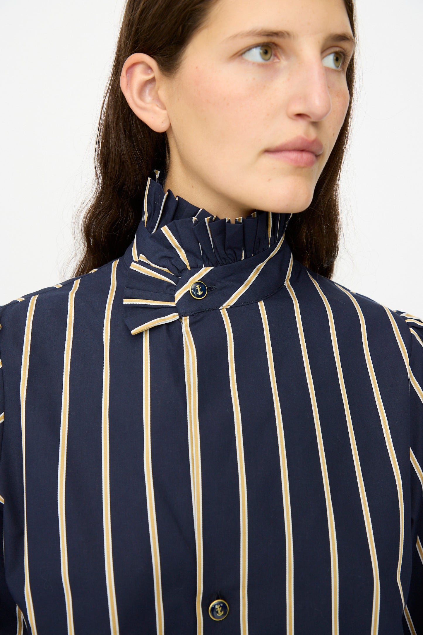 A person wears the Les Prairies De Paris Cotton Josephine Shirt in R Studio, featuring navy blue with beige vertical stripes and a ruffled high collar, looking to the side against a plain background.
