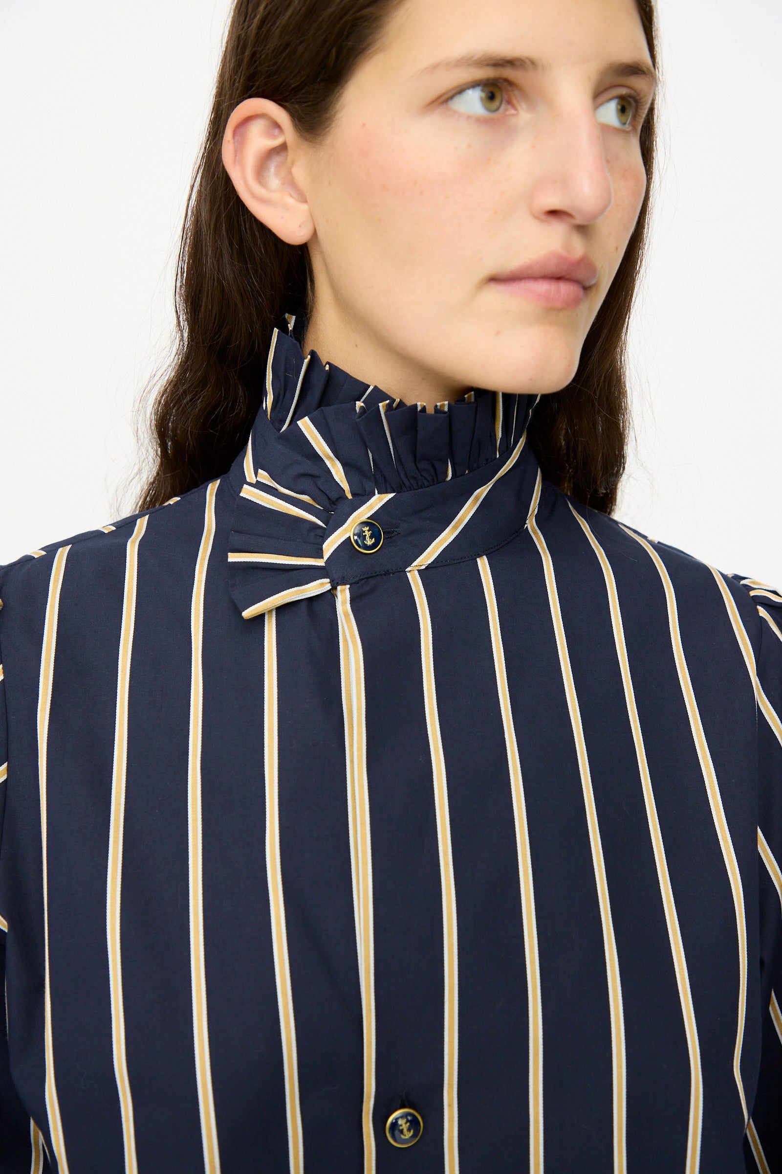 A person wears the Les Prairies De Paris Cotton Josephine Shirt in R Studio, featuring navy blue with beige vertical stripes and a ruffled high collar, looking to the side against a plain background.