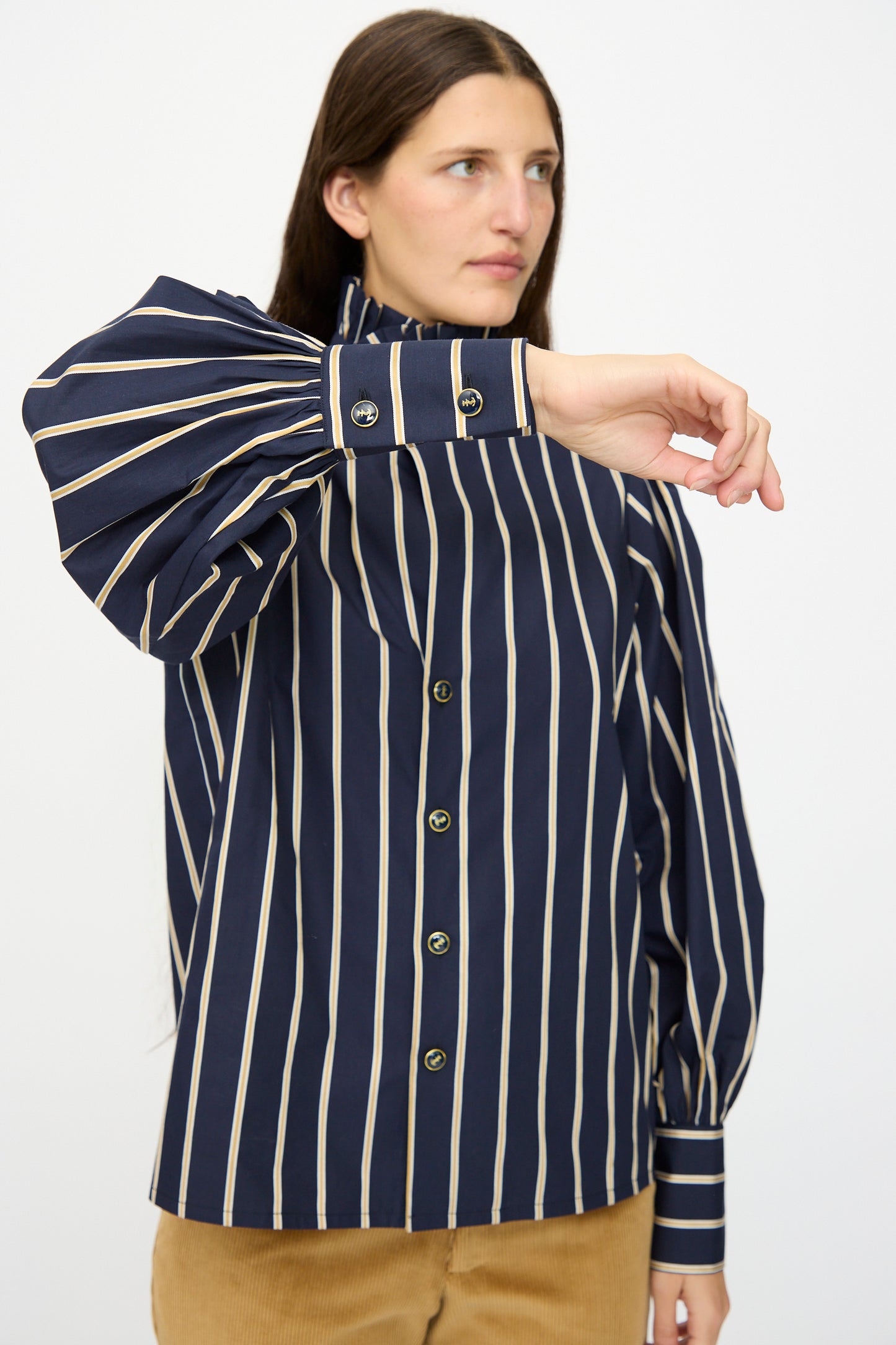 A person wearing the Les Prairies De Paris Cotton Josephine Shirt in R Studio, featuring navy blue and beige stripes with puff sleeves, paired with beige pants, raises one arm across their chest.