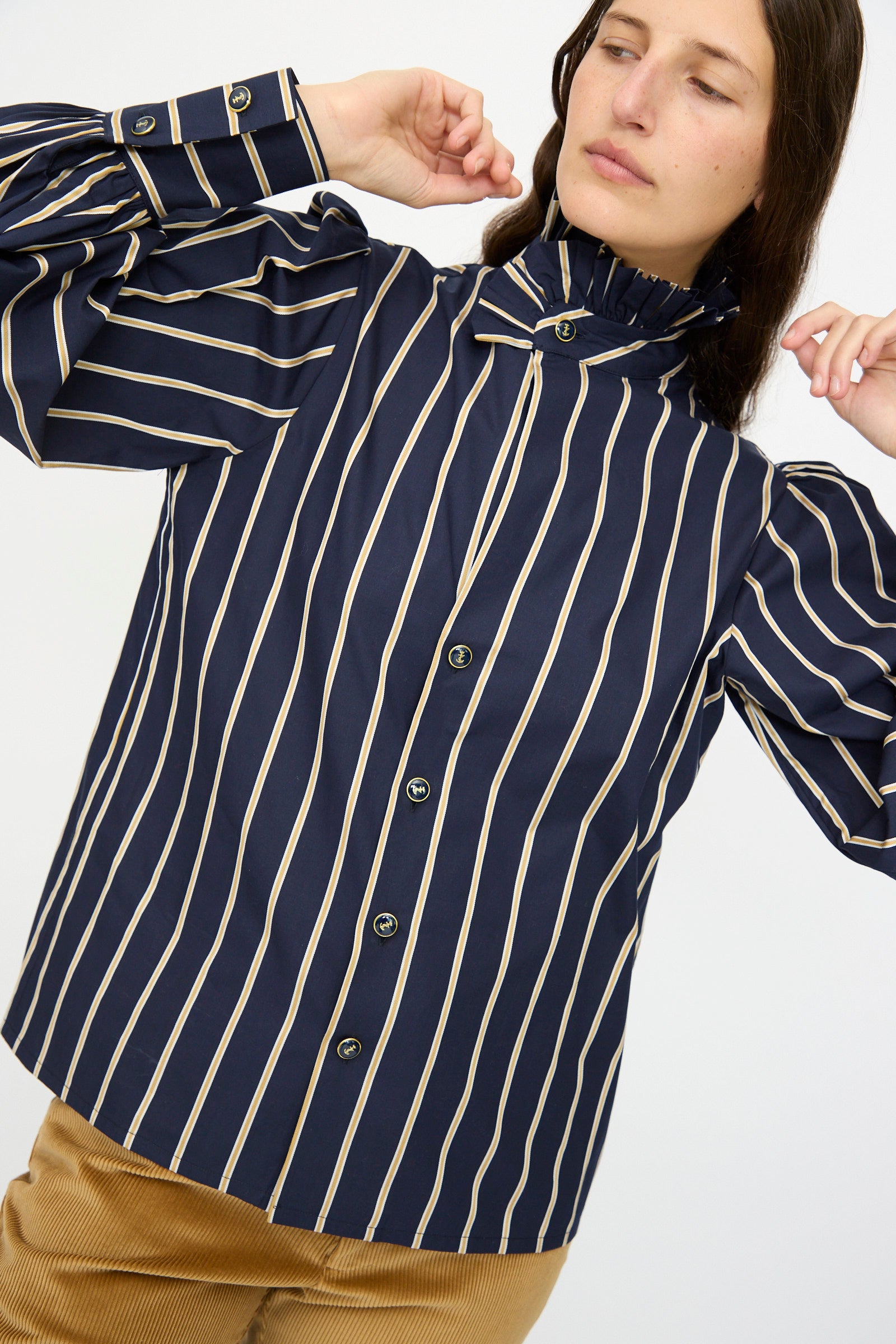 A person wears the Cotton Josephine Shirt in R Studio by Les Prairies De Paris, featuring navy blue and beige stripes, puff sleeves, and a ruffled collar, paired with tan pants.