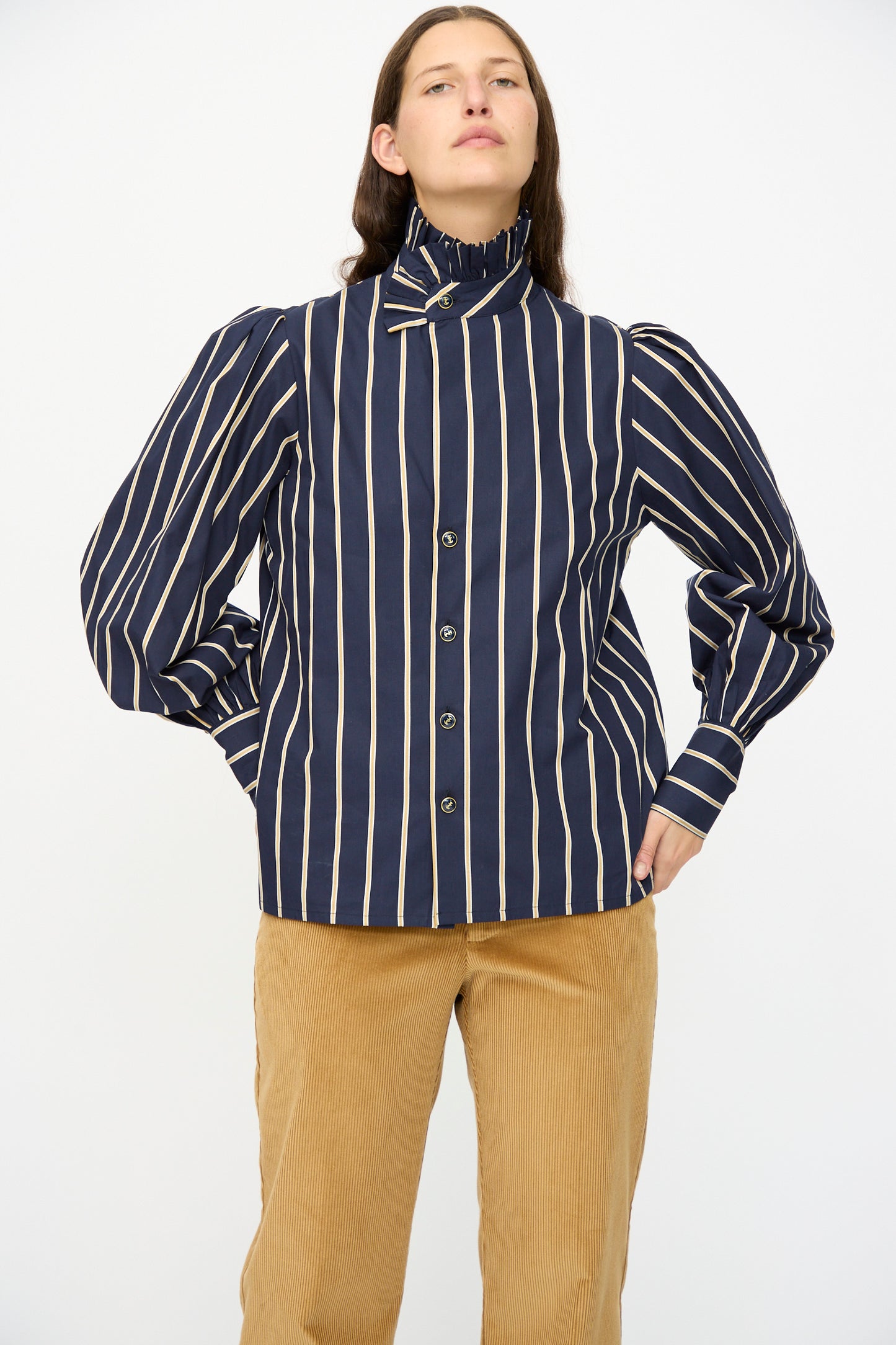 A person wears the Cotton Josephine Shirt in R Studio by Les Prairies De Paris—navy blue and white stripes with a ruffled collar and puffed sleeves—paired with tan corduroy pants, standing against a plain white background.