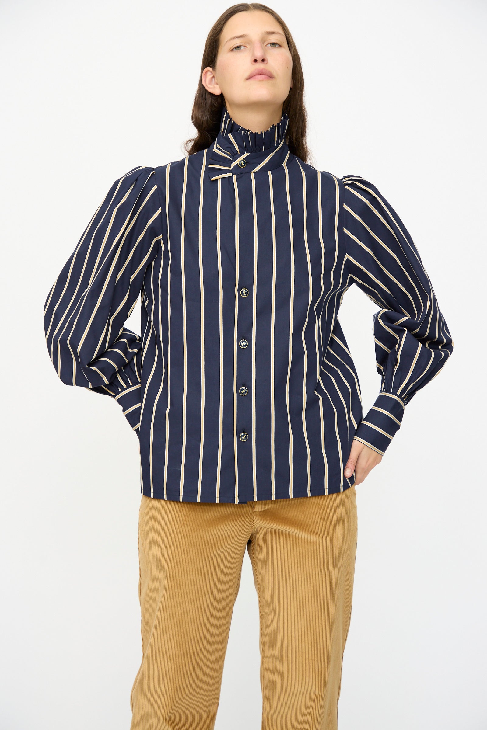 A person wears the Cotton Josephine Shirt in R Studio by Les Prairies De Paris—navy blue and white stripes with a ruffled collar and puffed sleeves—paired with tan corduroy pants, standing against a plain white background.