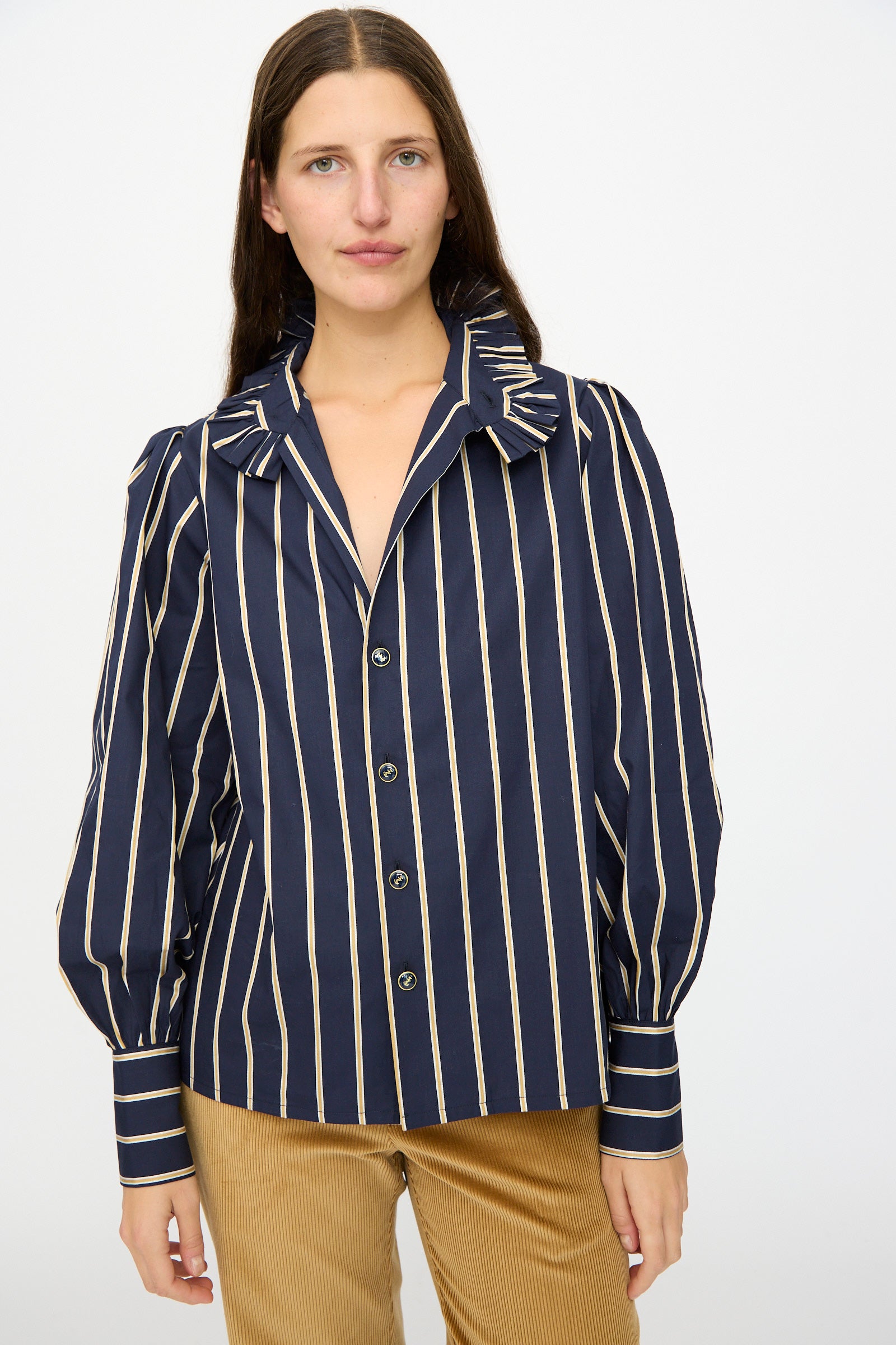 A woman wears the Les Prairies De Paris Cotton Josephine Shirt in R Studio, featuring navy blue stripes, puff sleeves, and a ruffled collar, paired with tan corduroy pants against a plain background.