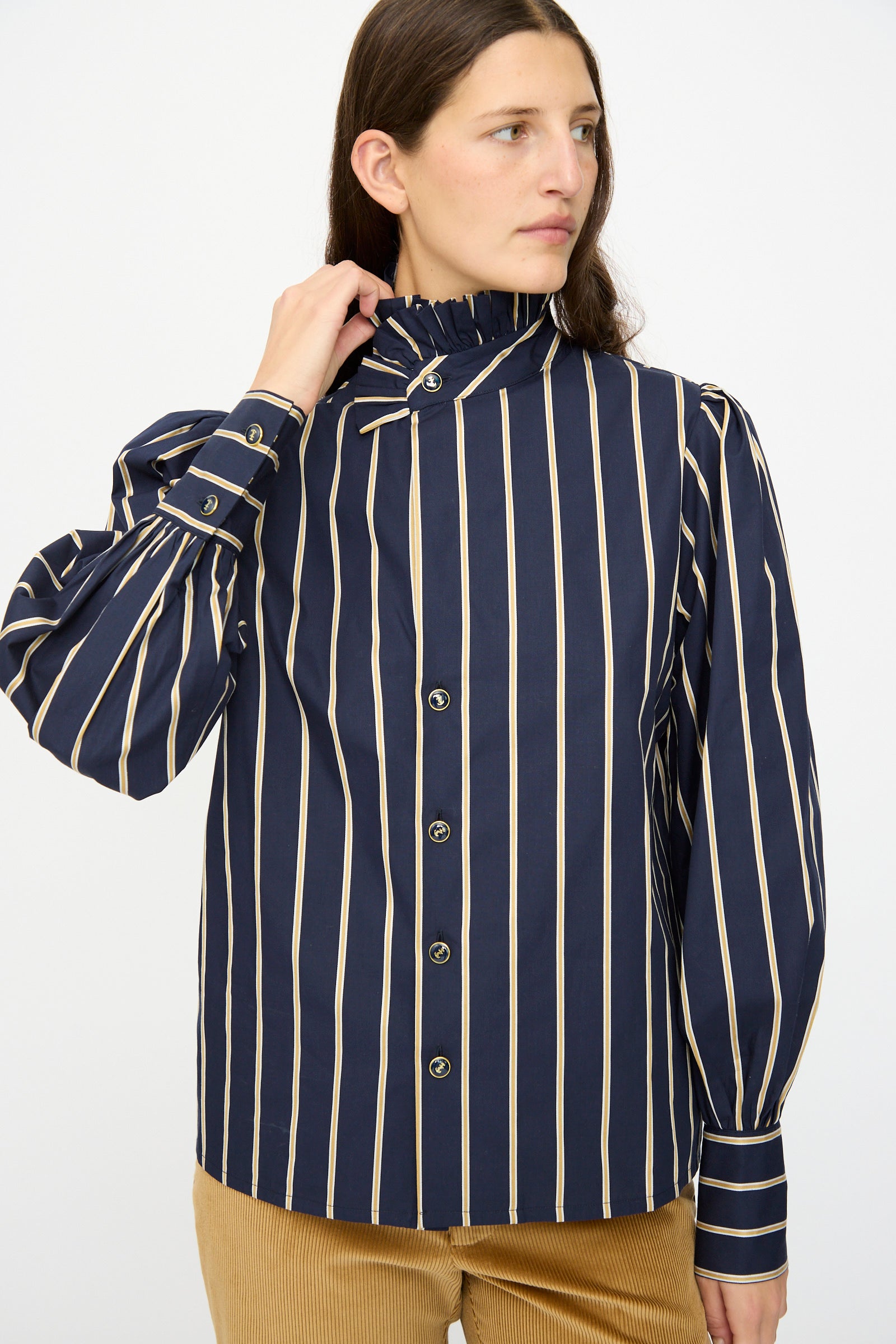 A person wears the Les Prairies De Paris Cotton Josephine Shirt in R Studio, featuring navy blue cotton with beige vertical stripes, puff sleeves, and a ruffled collar, standing against a plain background.