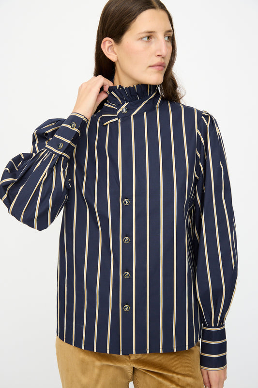 A person wears the Les Prairies De Paris Cotton Josephine Shirt in R Studio, featuring navy blue cotton with beige vertical stripes, puff sleeves, and a ruffled collar, standing against a plain background.