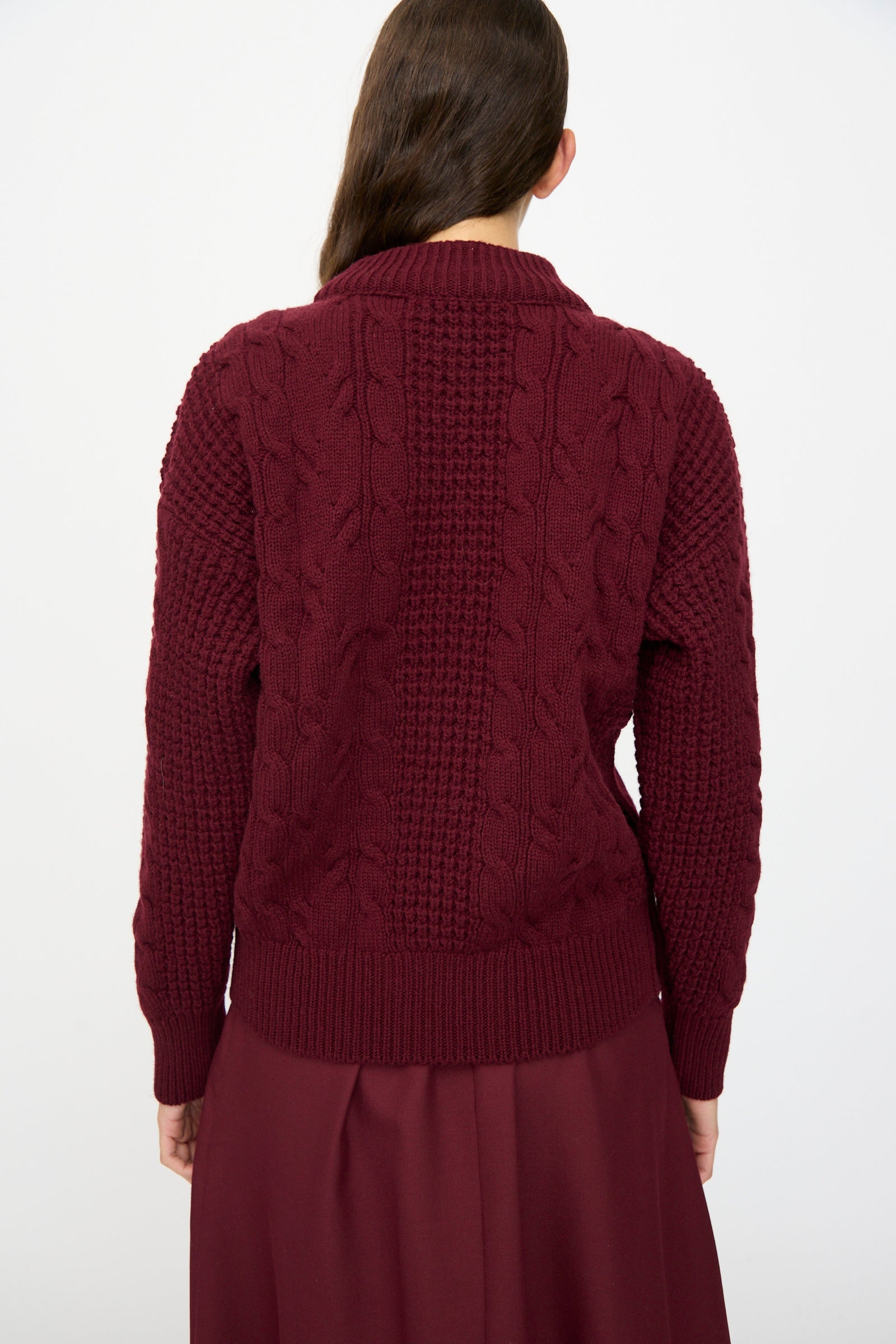 A person with long brown hair is shown from the back against a white background, wearing Les Prairies De Paris's The Irish Wool Sweater in Bordeaux with a matching skirt.