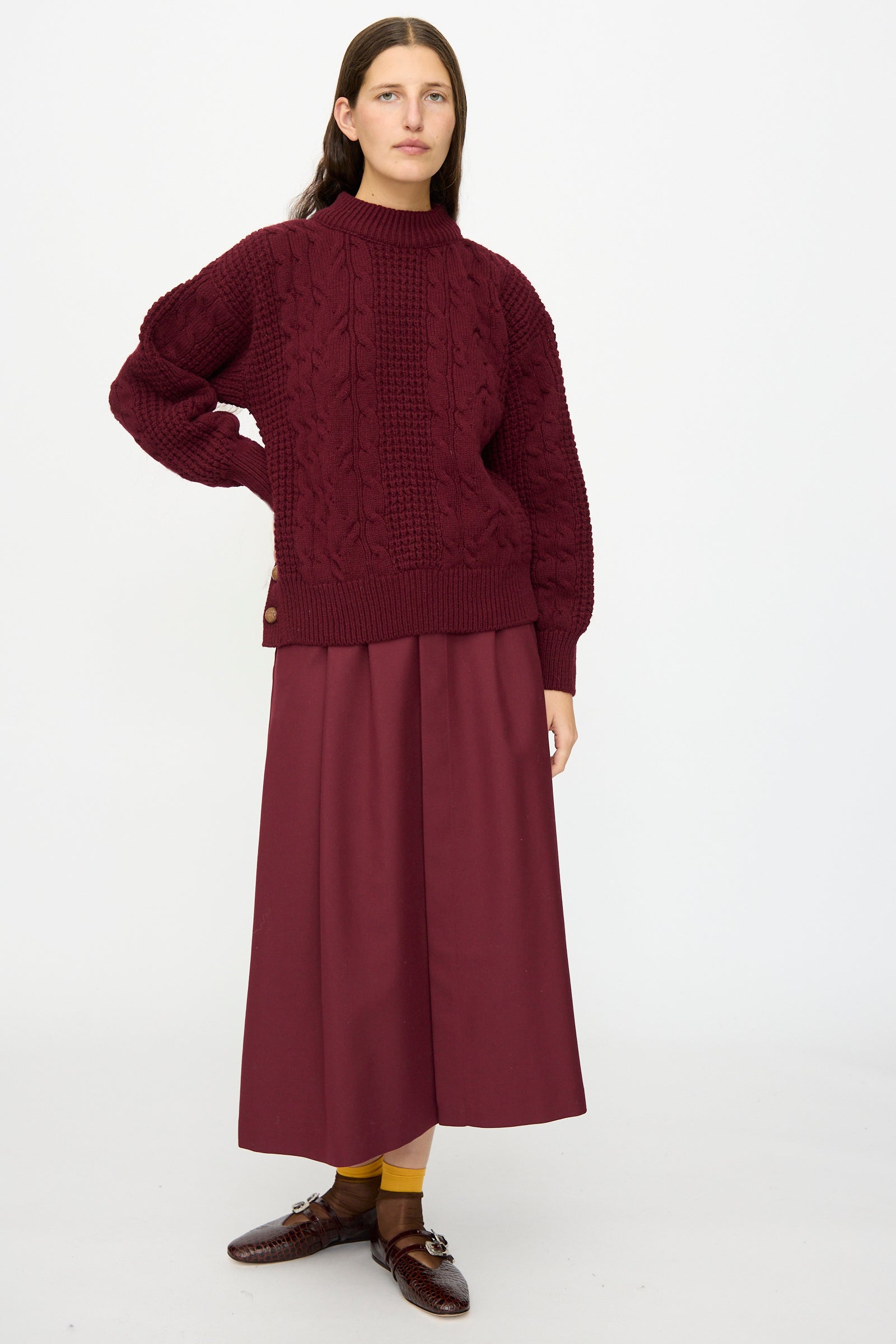 A person stands against a white background wearing Les Prairies De Paris's The Irish Wool Sweater in Bordeaux, paired with a matching skirt, brown shoes, and yellow socks.