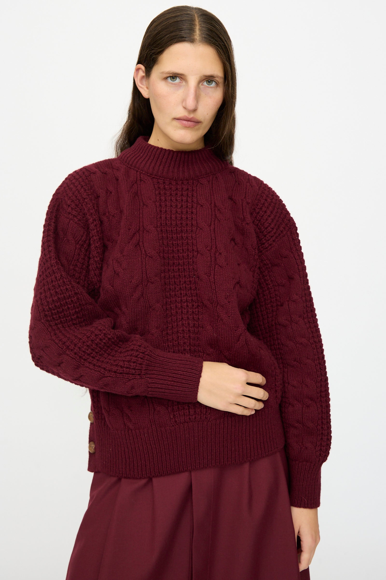 A person with long brown hair wears Les Prairies De Paris's The Irish Wool Sweater in Bordeaux with a matching skirt, standing against a plain white background.