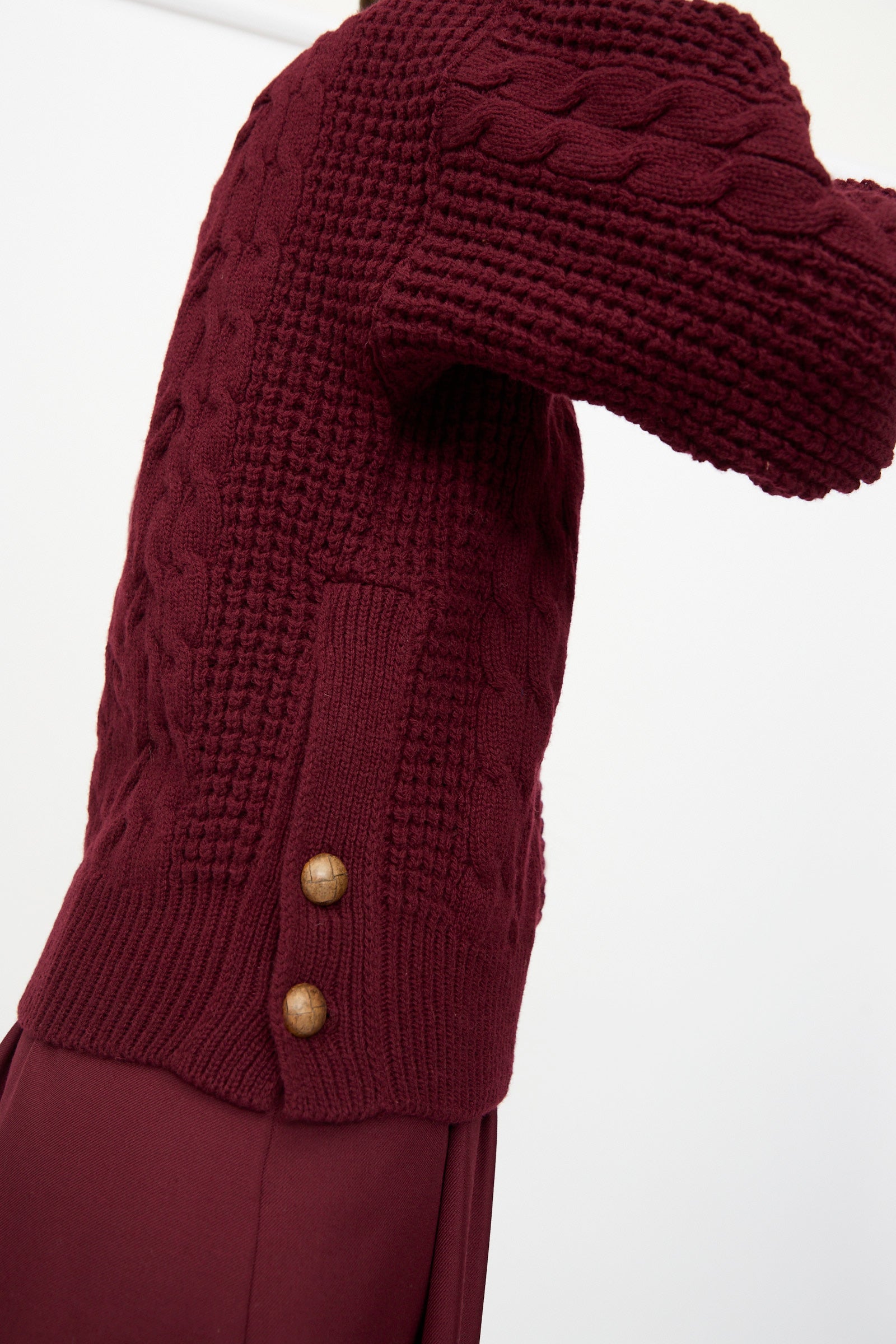 A close-up of Les Prairies De Paris' The Irish Wool Sweater in Bordeaux—a French-made, chunky cable knit wool pullover with textured sleeves and two brown side buttons—worn over a matching burgundy skirt.