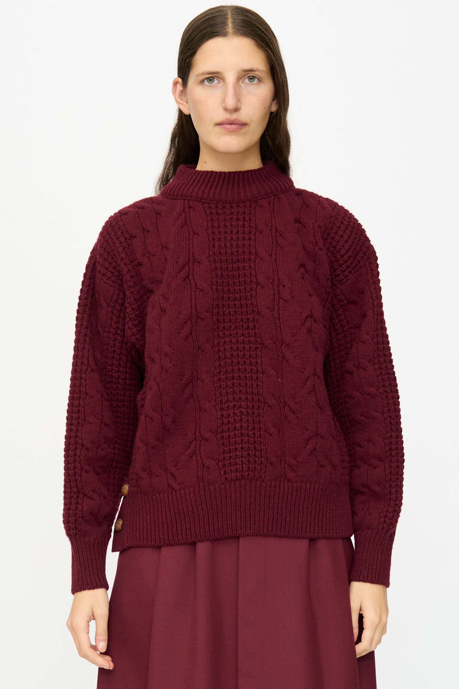 A person wearing The Irish Wool Sweater in Bordeaux by Les Prairies De Paris, featuring a cable knit design and long sleeves, paired with a matching skirt, stands against a plain white background.