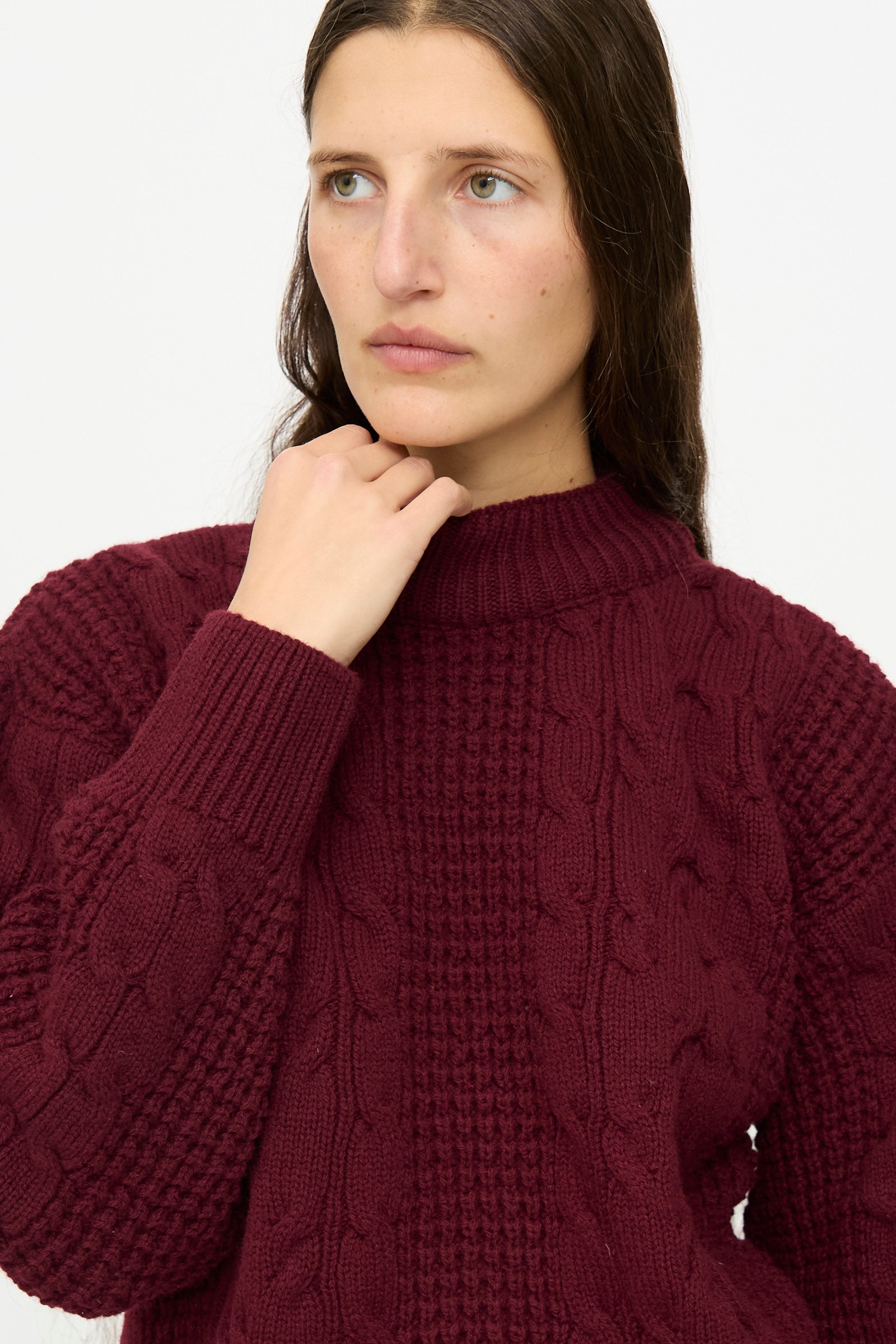 A person with long brown hair wears The Irish Wool Sweater in Bordeaux by Les Prairies De Paris, posing with one hand near their chin against a plain light background.